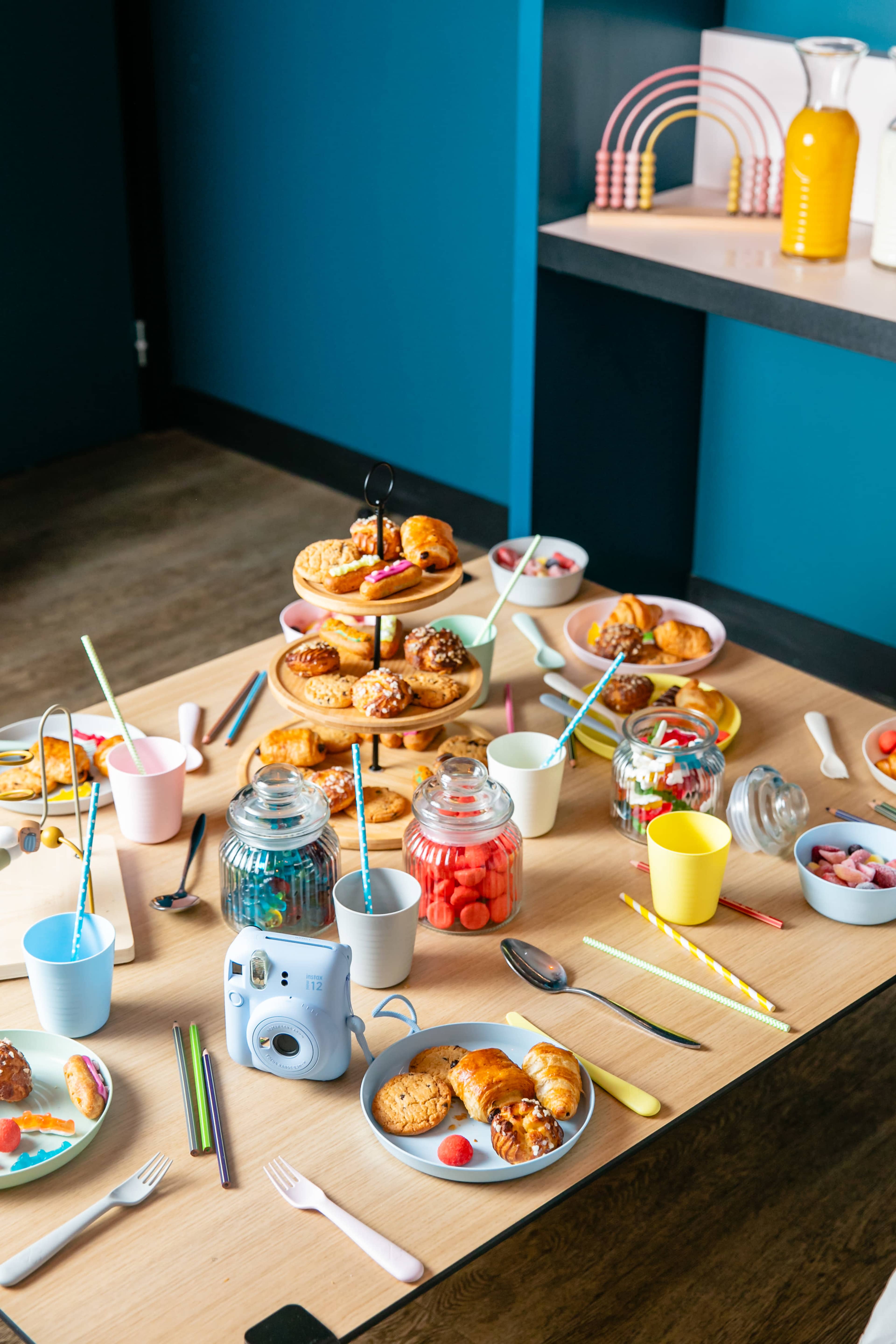 A tabletop is set with a variety of colorful pastries, candies, and beverages, surrounded by plates, cups, and a camera.