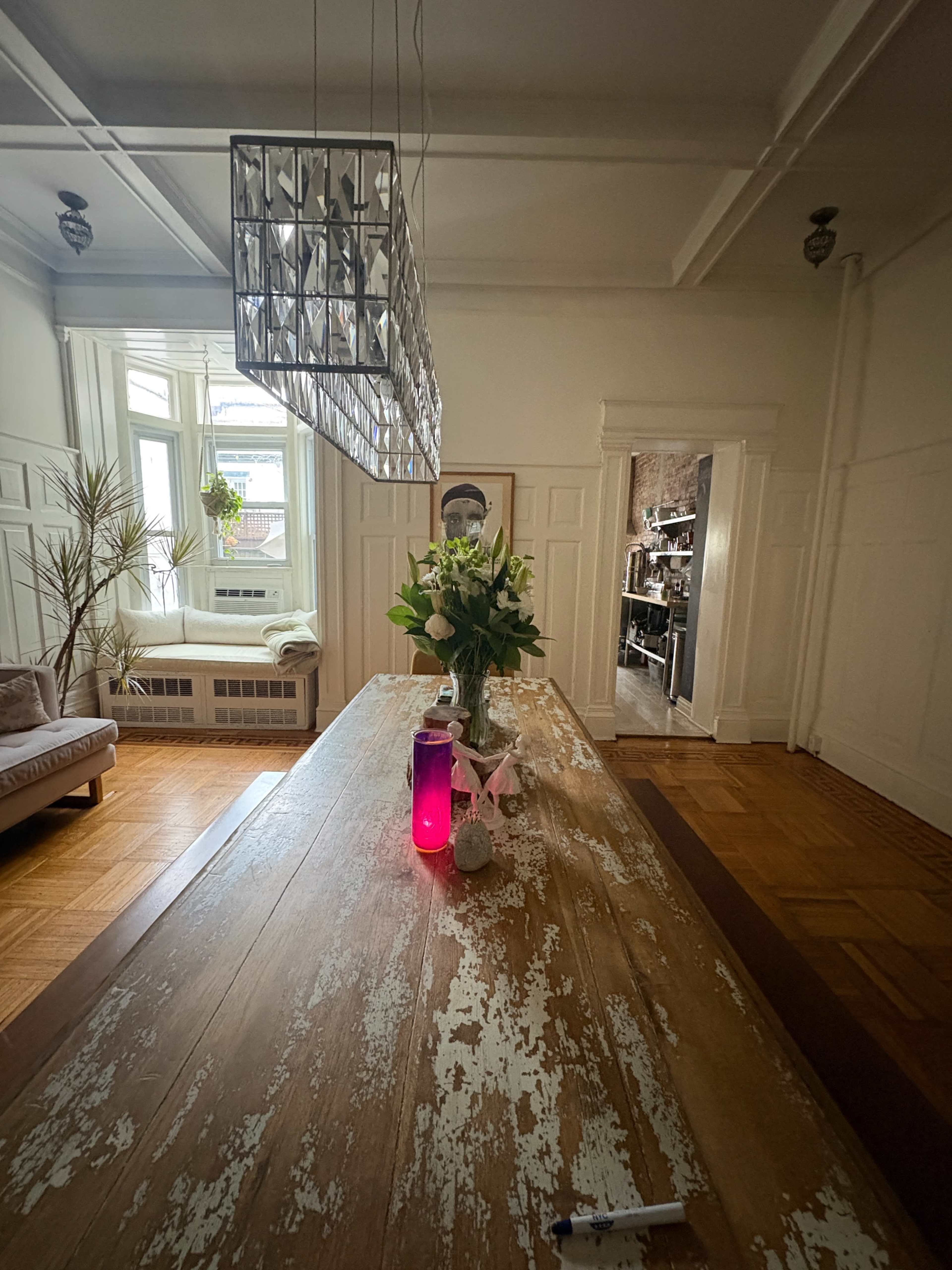A long wooden dining table with a pink water bottle and a vase of flowers is illuminated by a modern chandelier in a bright, spacious room leading to a kitchen area.