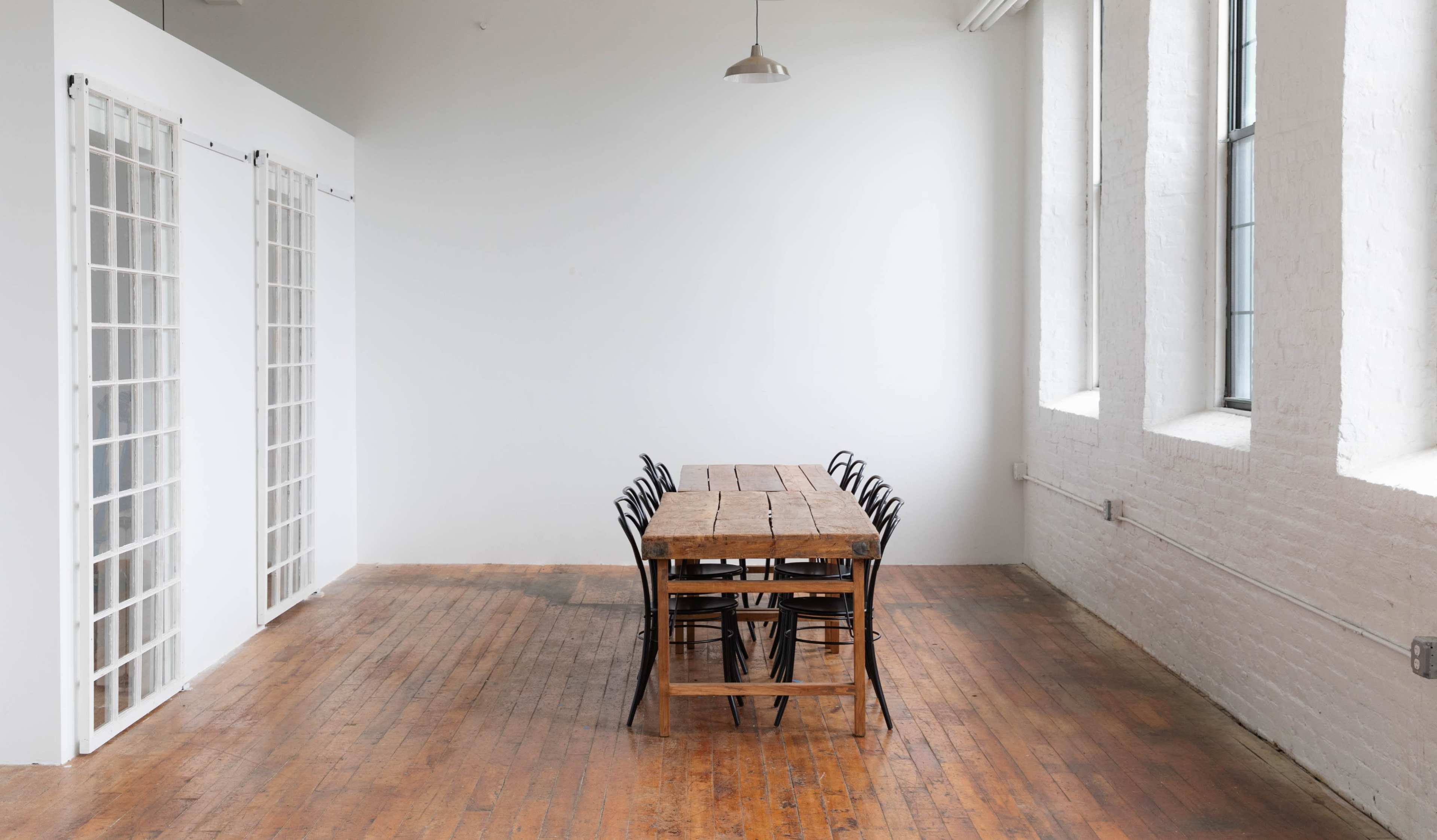 A long wooden table with black chairs is positioned in an empty, brightly lit room with white walls and large windows.