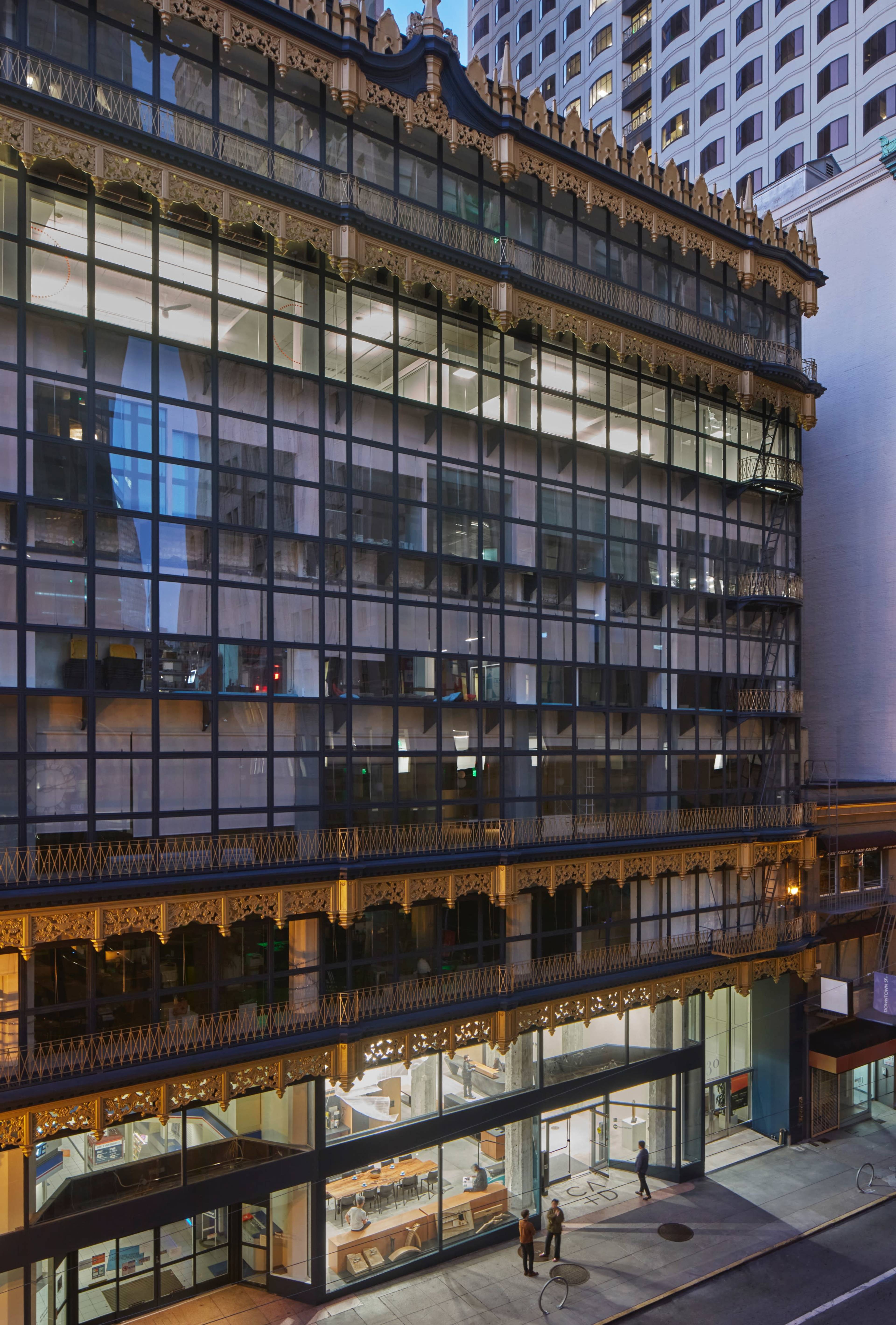 A modern glass building with ornate detailing, situated in an urban environment, where people walk past the entrance at dusk.