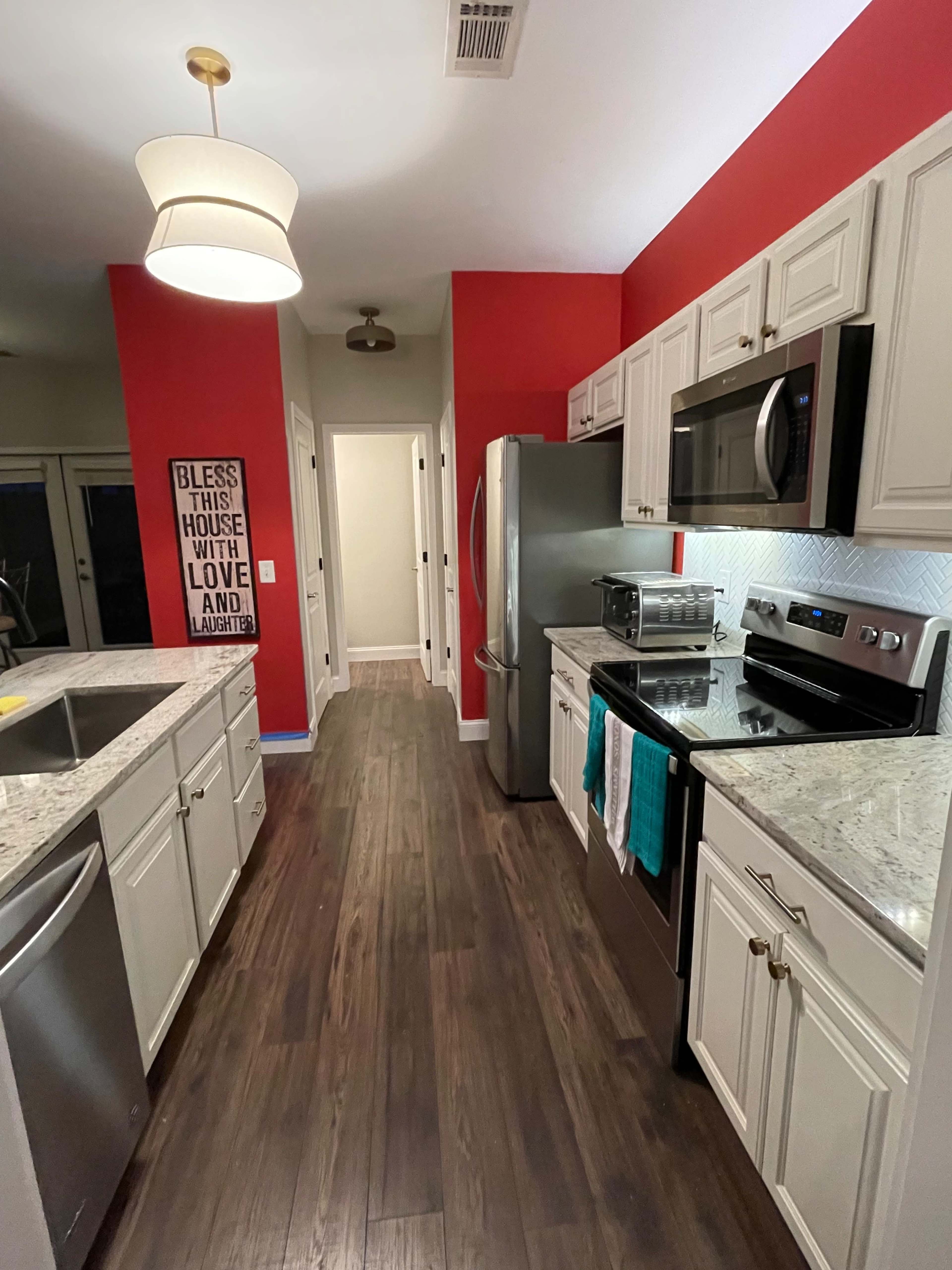 A modern kitchen features red accent walls, stainless steel appliances, and granite countertops, with a pendant light overhead.