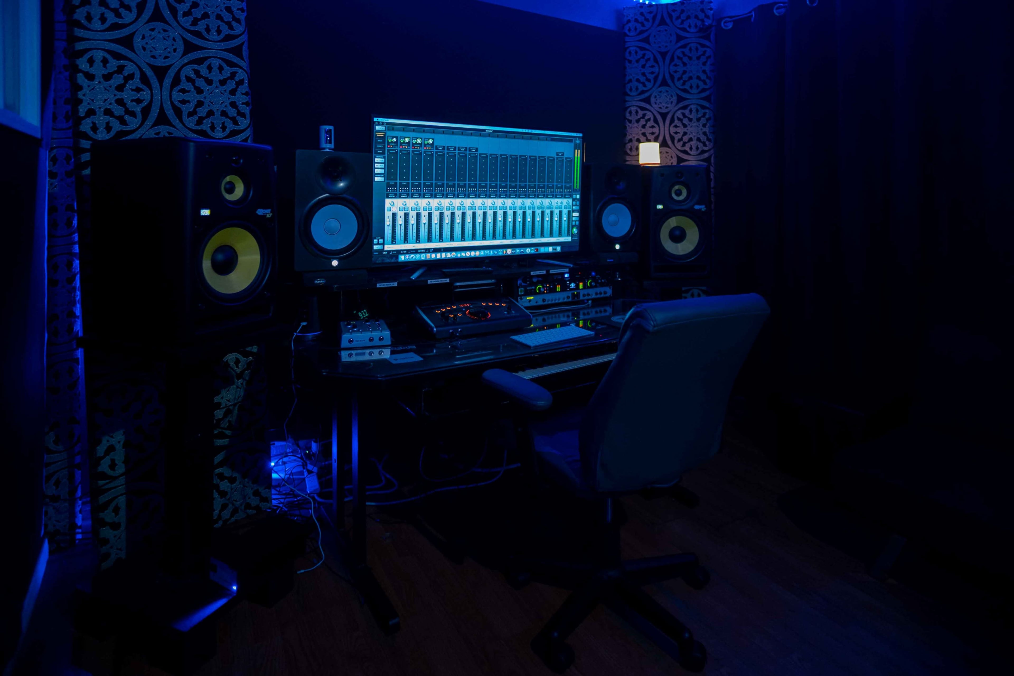 A music production studio features a computer monitor displaying audio mixing software, flanked by two speaker monitors and a black office chair in a dimly lit environment.