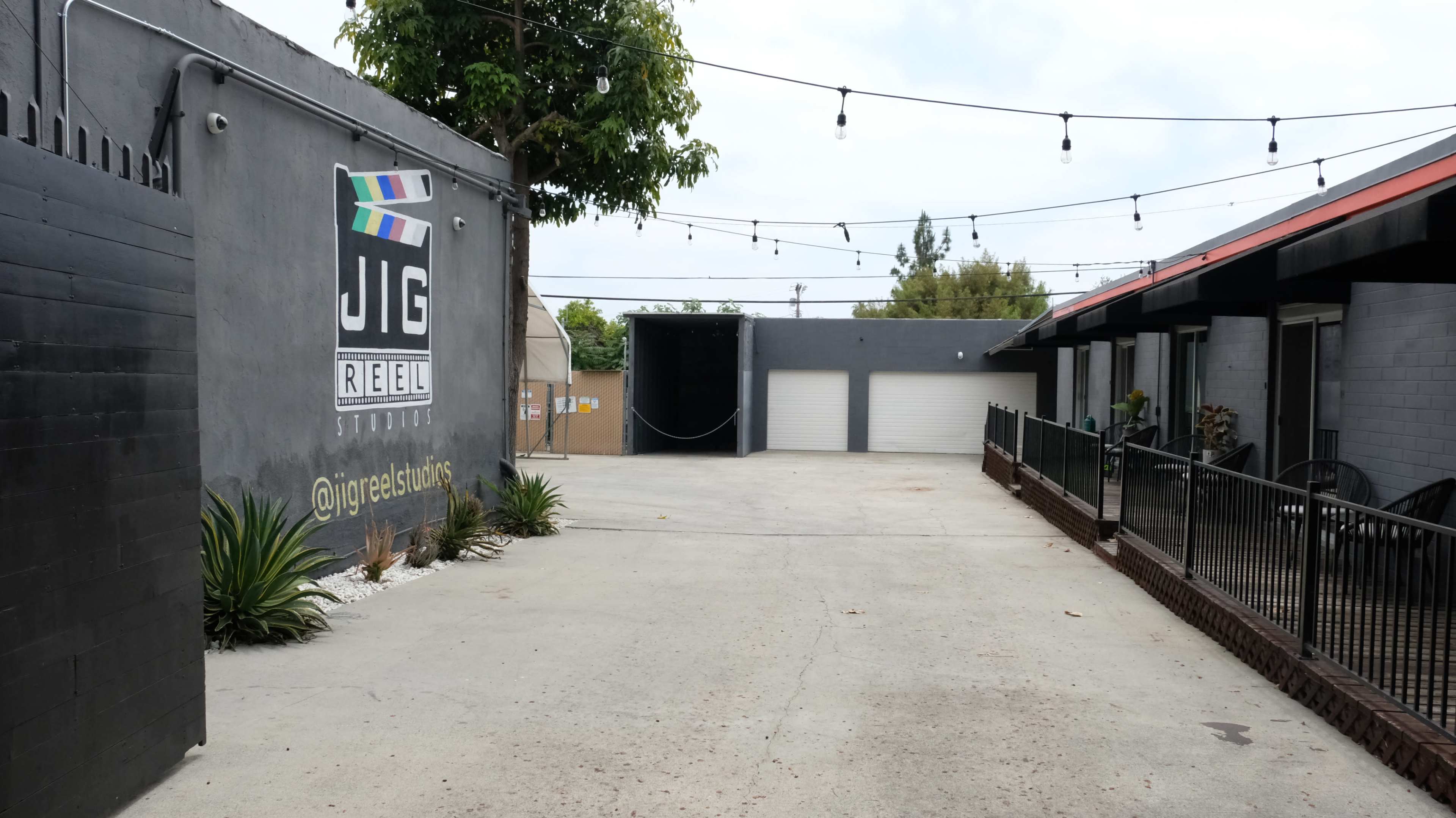 The image shows a courtyard with a gray wall featuring a sign for "JIG REEL STUDIOS" and string lights overhead, leading to a series of garages and buildings.