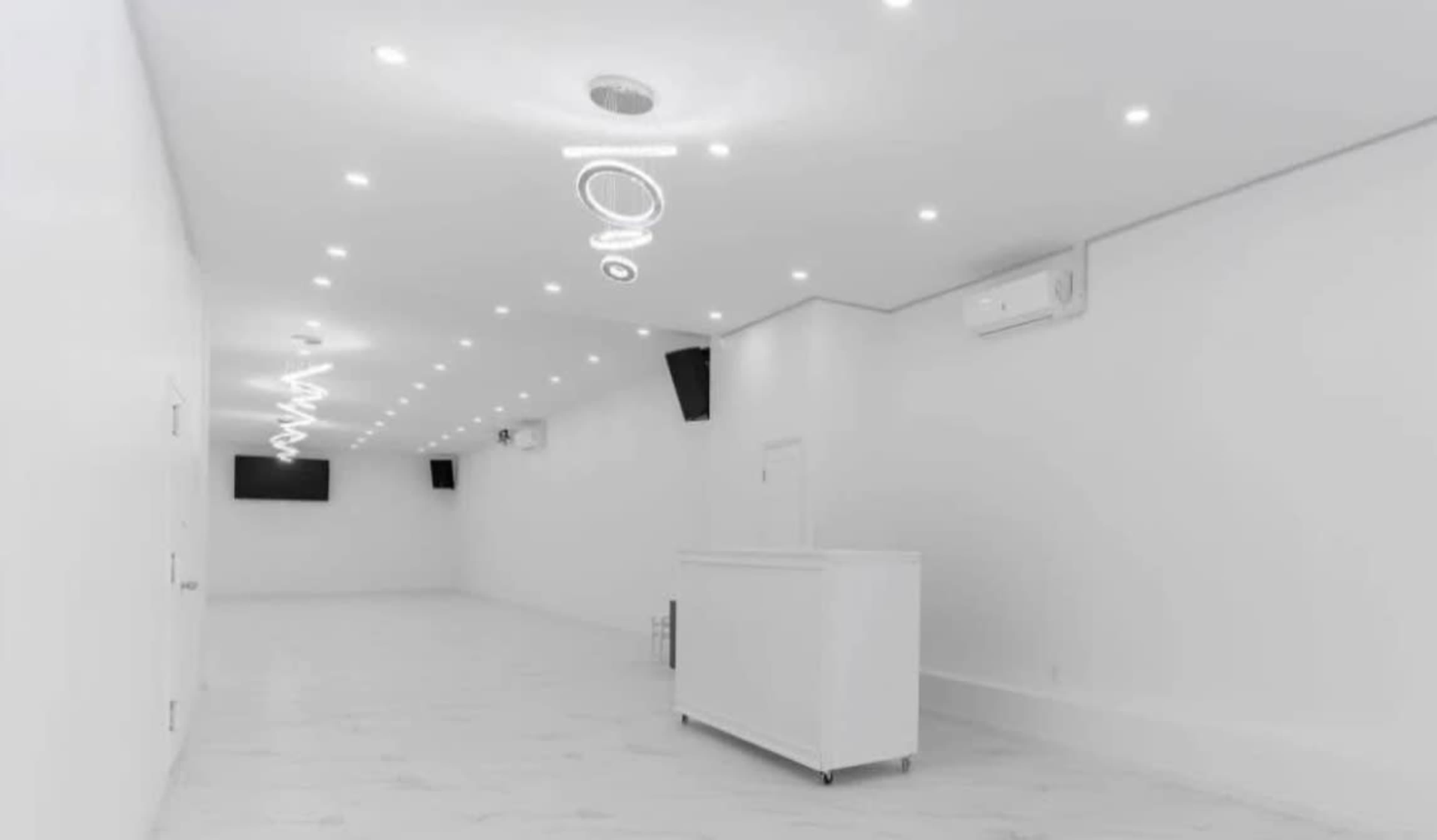 The image shows a spacious, white room with modern lighting, ceiling-mounted speakers, and a reception area at one end.