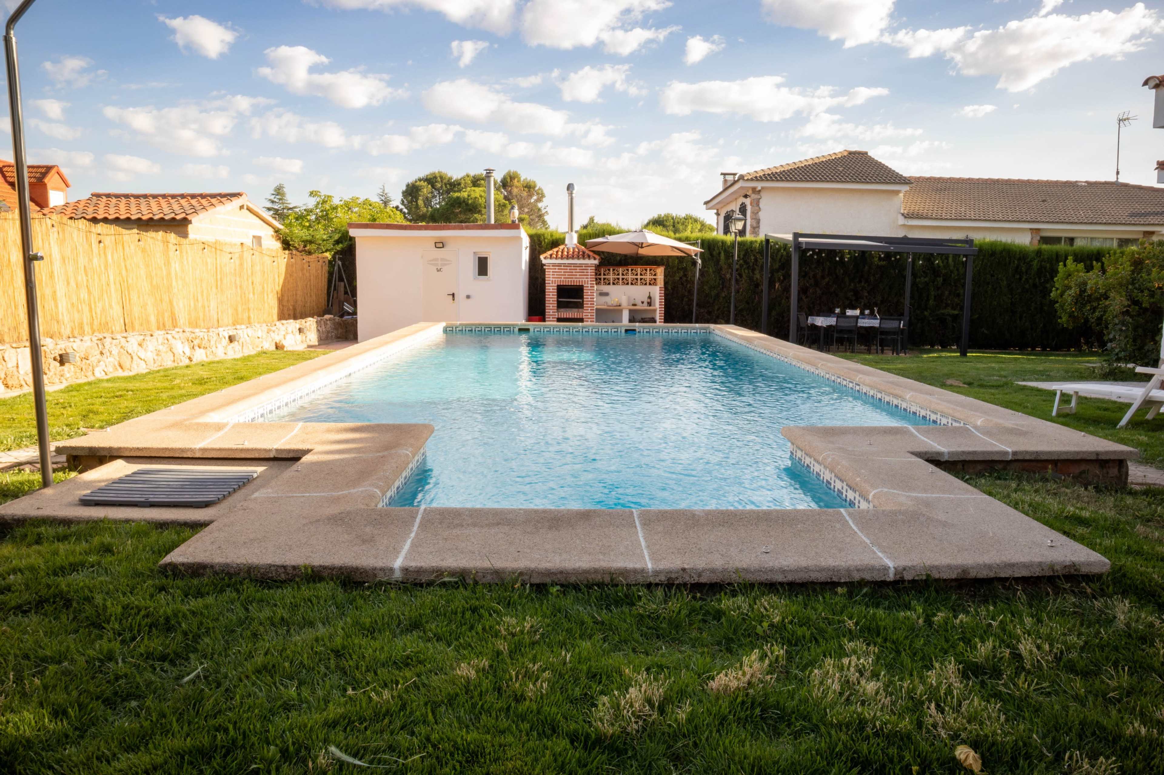 The image shows a rectangular swimming pool surrounded by grass, with a small structure and a barbecue area visible nearby.