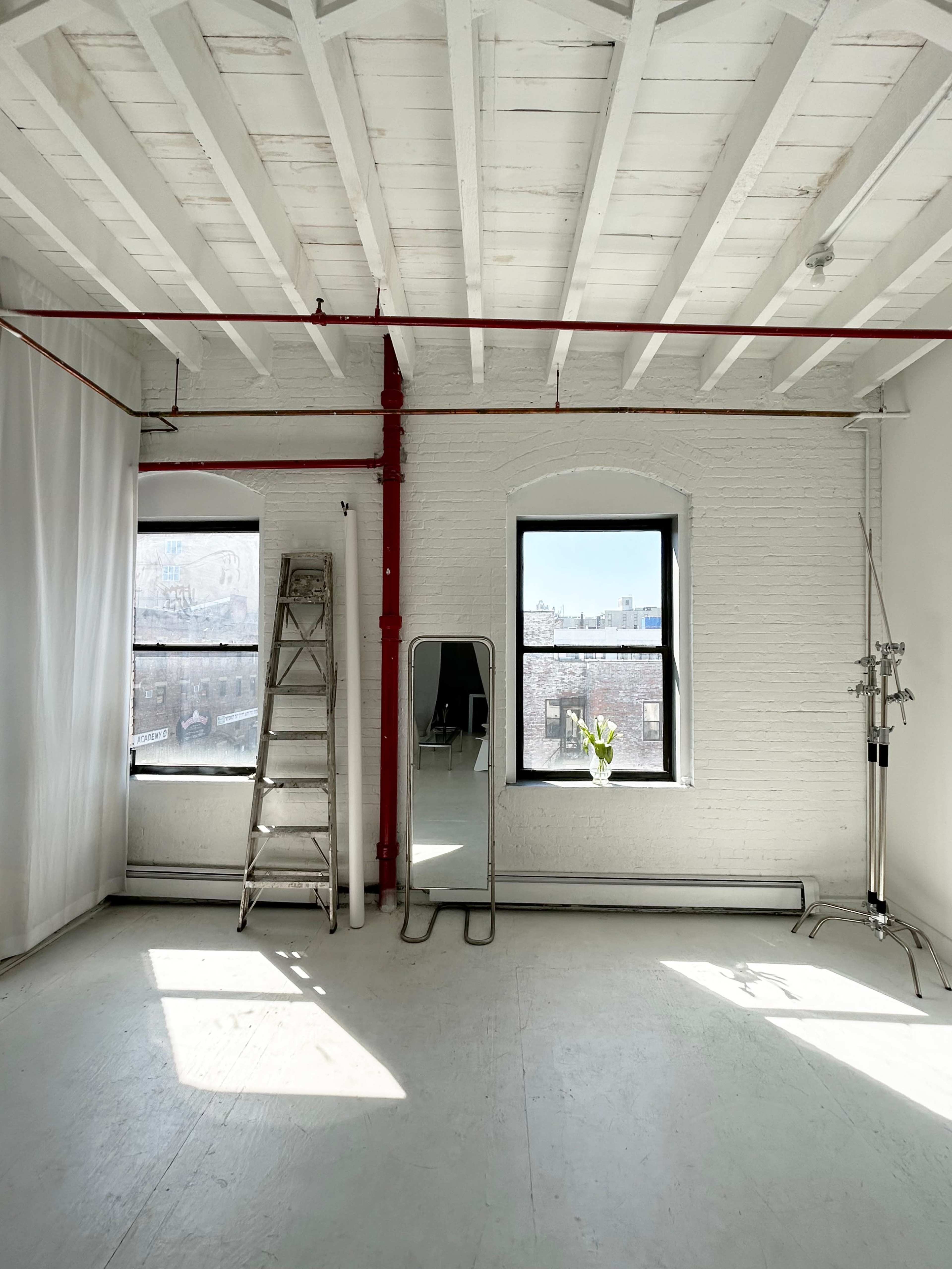 A bright, minimalist room with white walls, a wooden ceiling, a ladder, a mirror, a metal stand, and two windows letting in natural light.