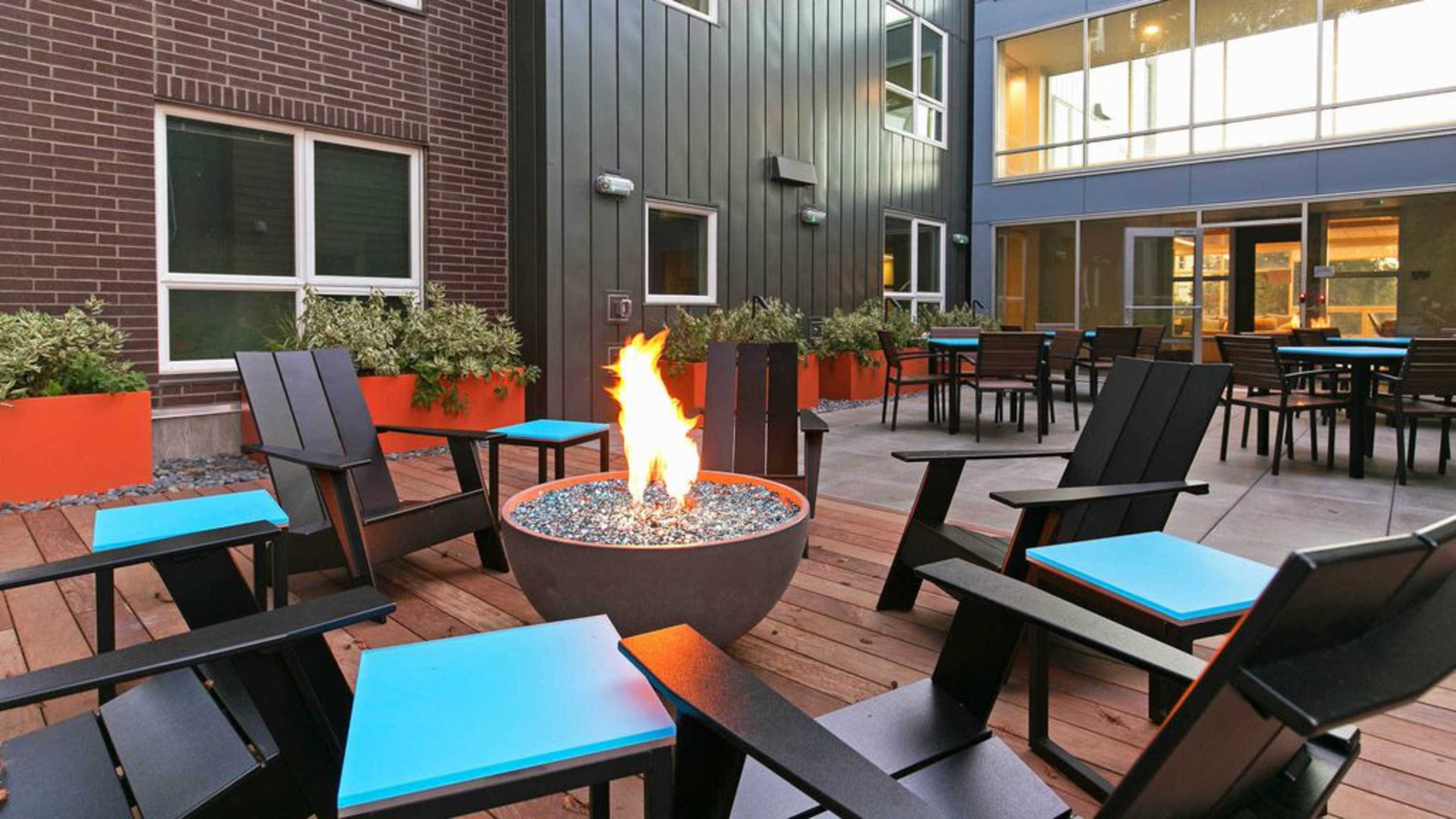 A courtyard with black chairs featuring blue accents arranged around a circular fire pit, surrounded by orange planters and large windows of a building.