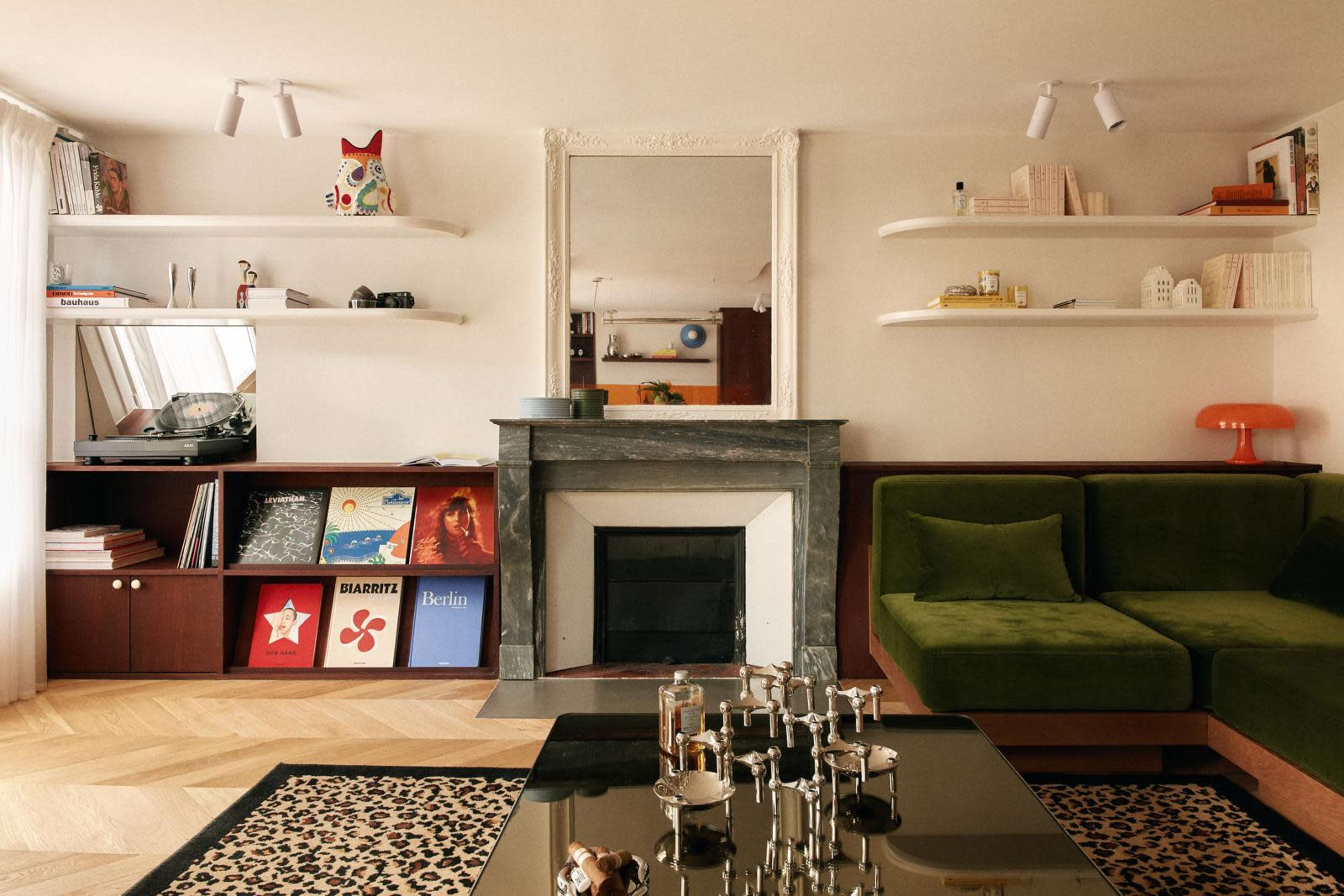 The image depicts a modern living room featuring a green velvet sofa, a marble fireplace, and various shelves displaying books and decorative items.