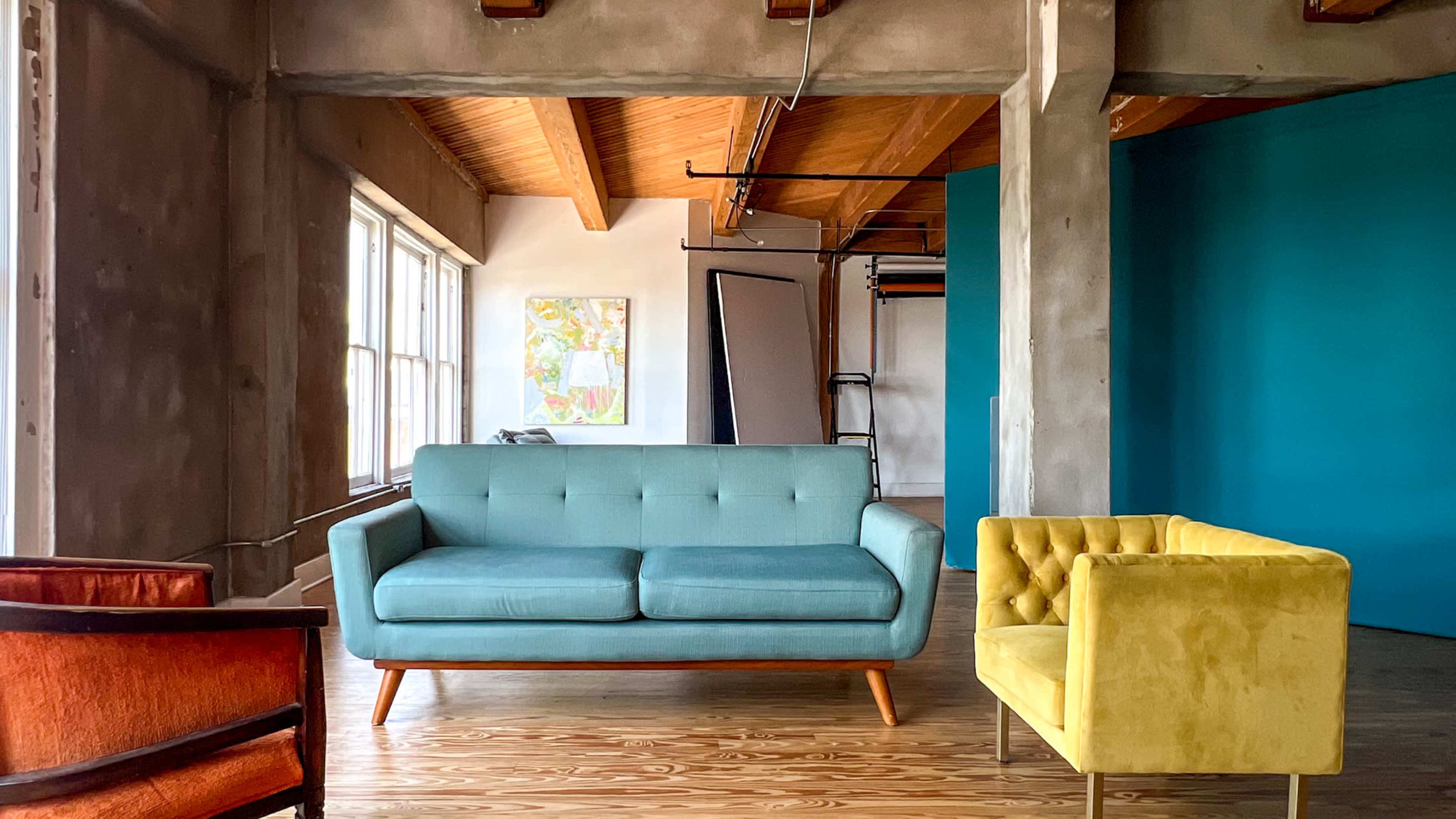 A spacious room featuring a light blue couch, a yellow armchair, and an orange armchair, with large windows and a teal wall in the background.