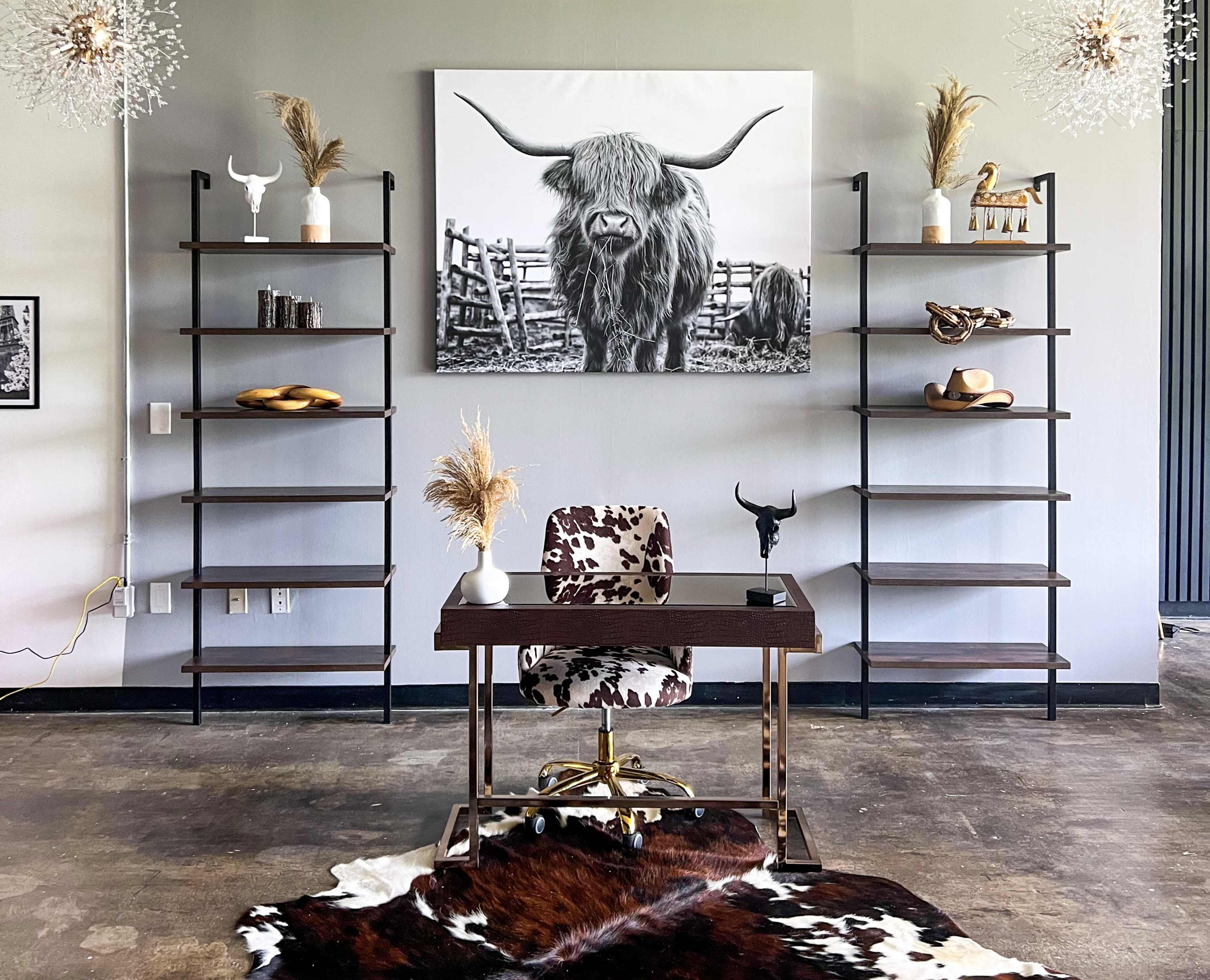 A modern workspace features a desk with a cowhide chair, surrounded by open shelving displaying decorative items and a large black-and-white photo of a highland cow on the wall.