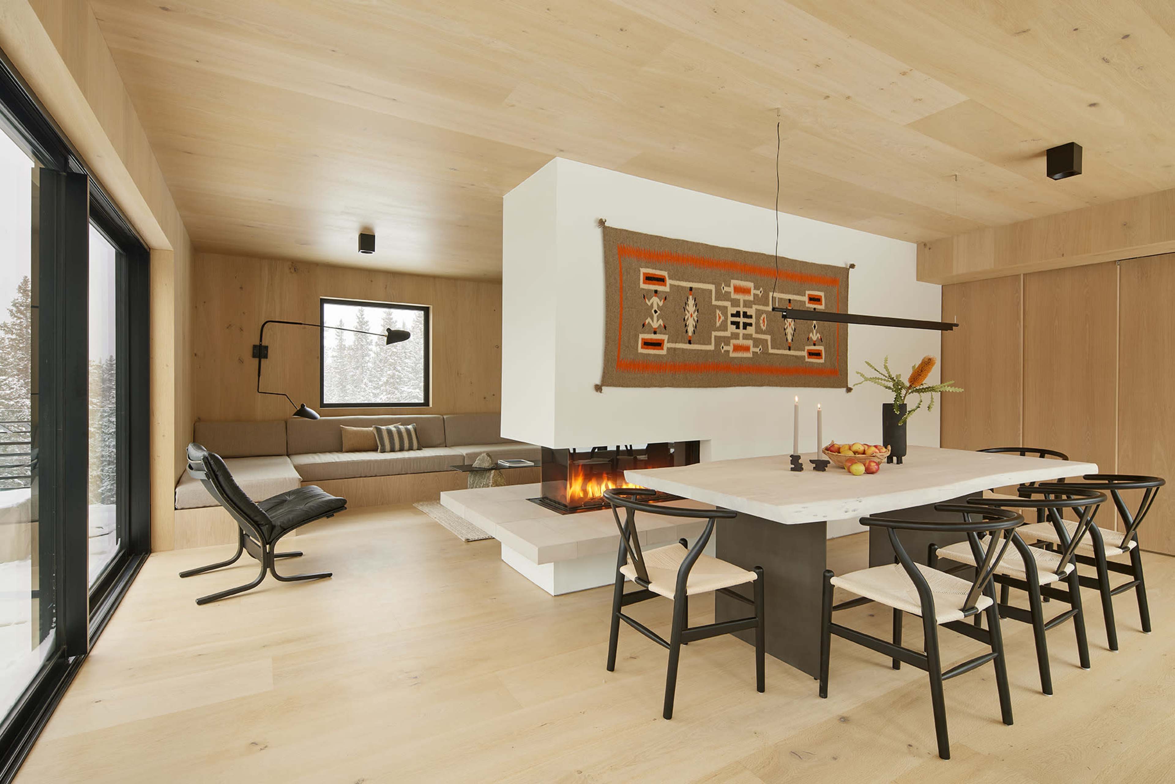 The image shows a modern living space featuring a minimalist dining area with a white table and black chairs, a cozy seating nook, and a fireplace, complemented by wood paneling and large windows.