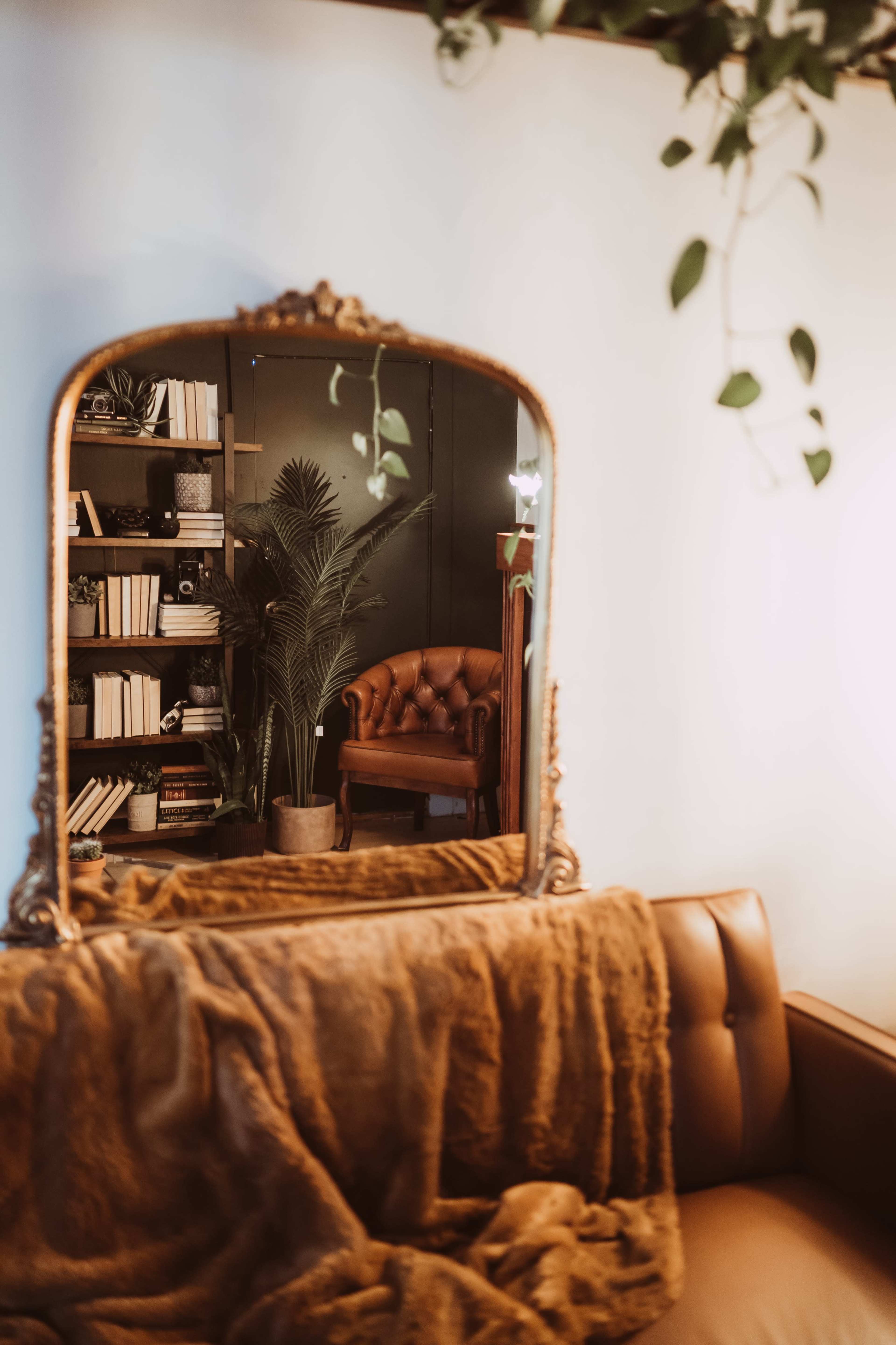 A large ornate mirror reflects a cozy corner of a room featuring a brown leather chair, bookshelves filled with books, and a potted plant.