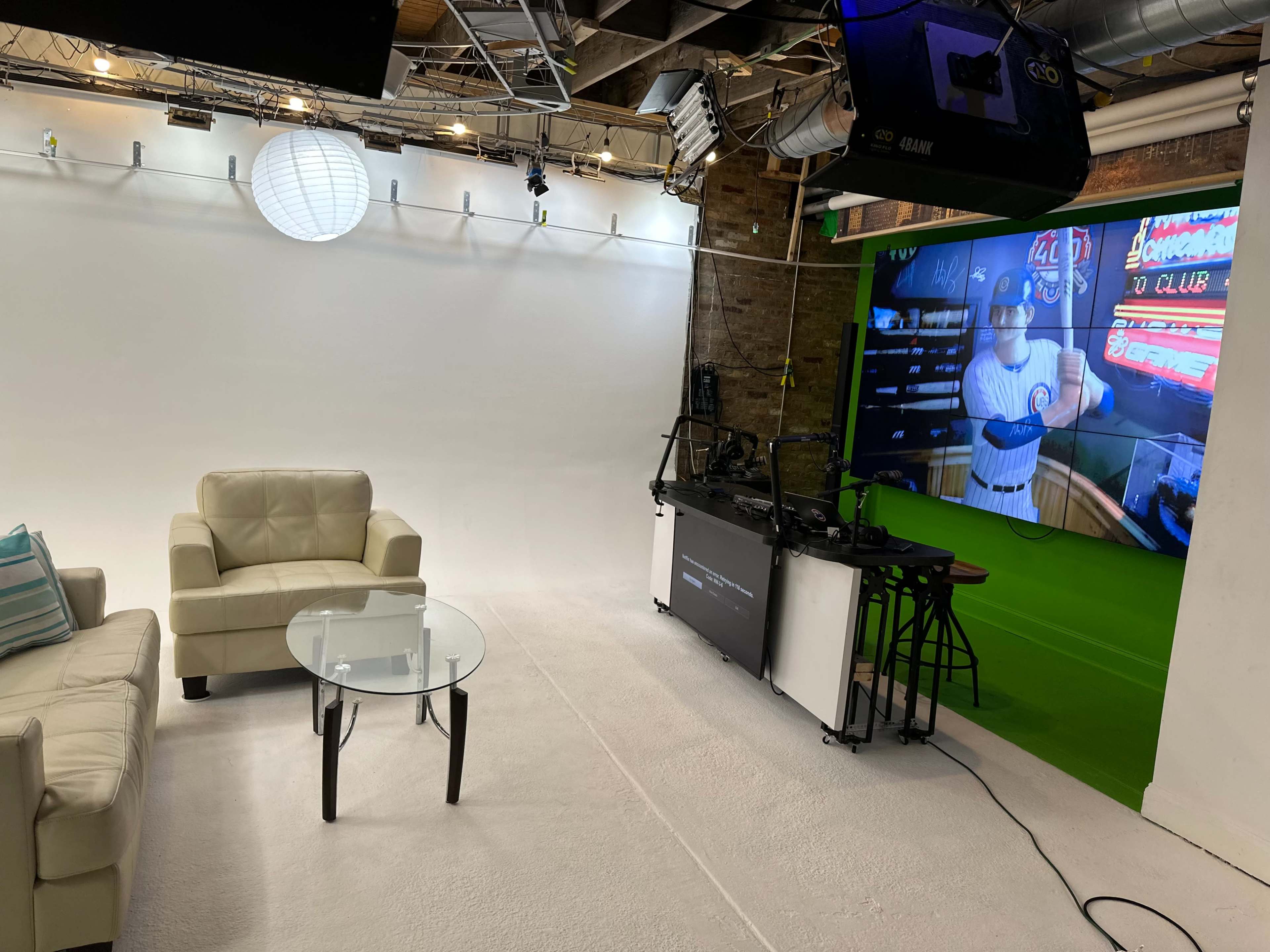 A small studio setup featuring a white backdrop, a light fixture, a couch, a glass coffee table, and a large screen displaying a baseball-themed scene.
