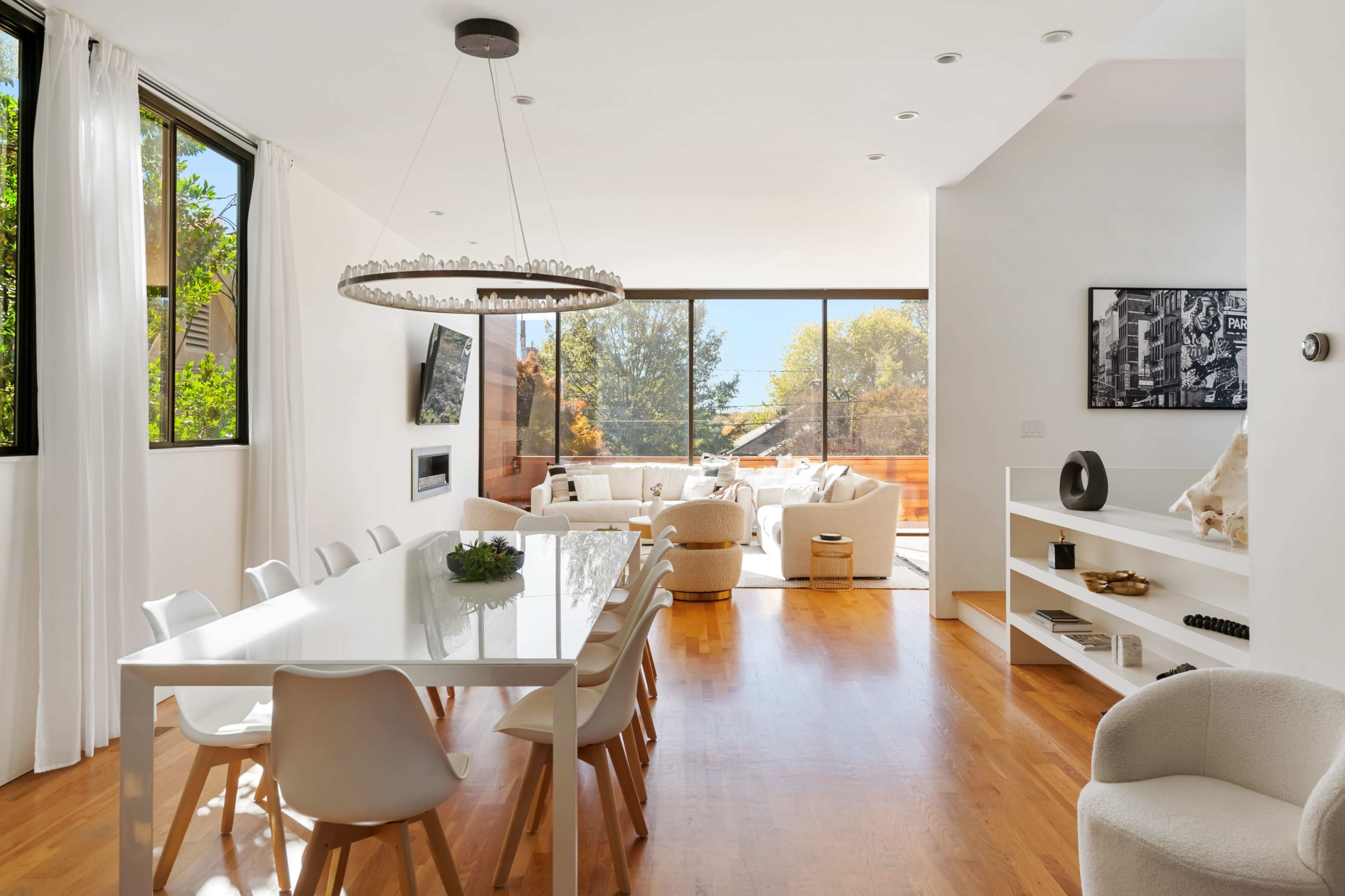 A bright and modern dining area features a long white table surrounded by chairs, leading into a spacious living room with large windows and contemporary furnishings.