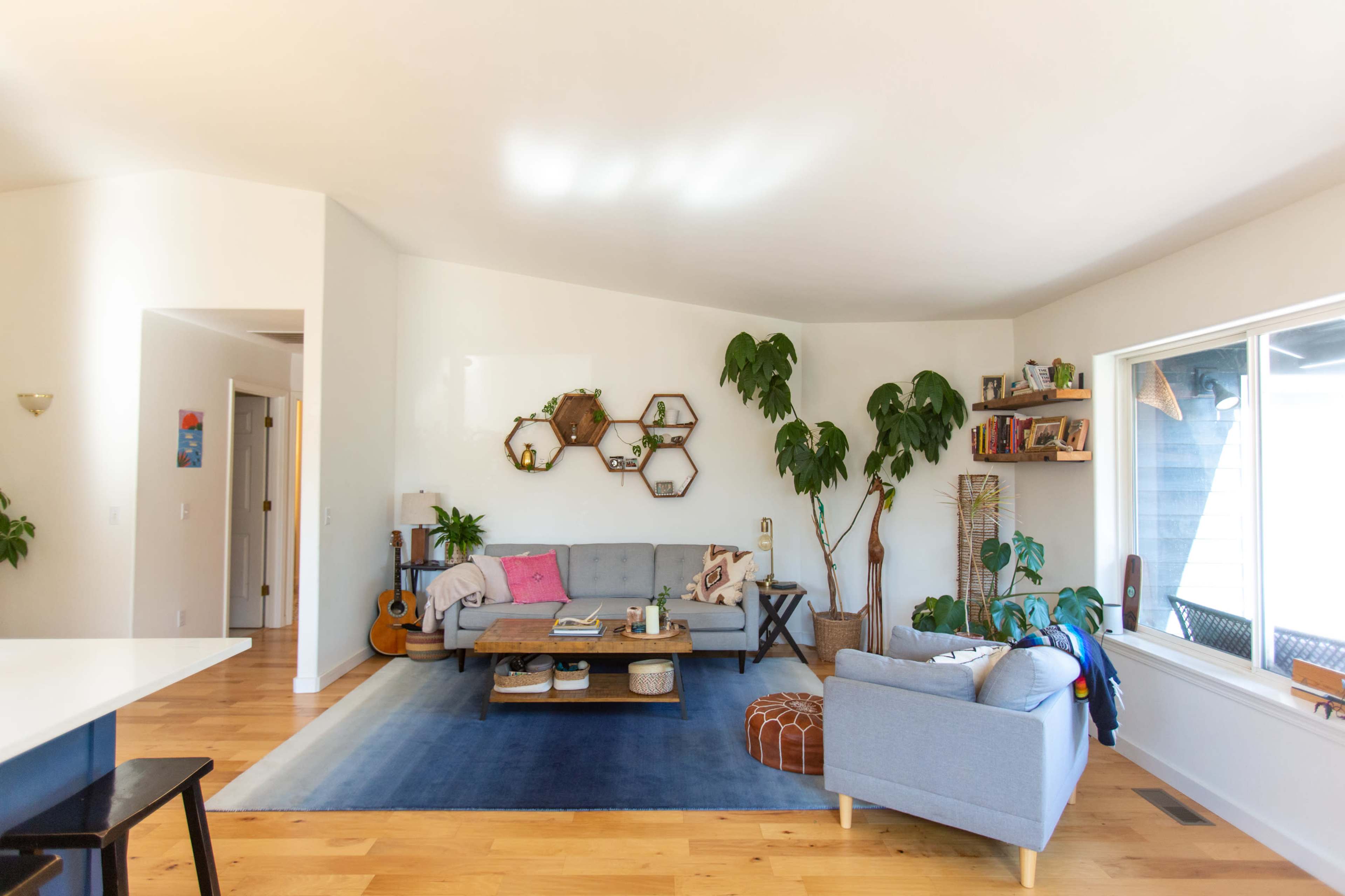 A spacious living room with a gray sofa, plants, a wooden coffee table, and a large window letting in natural light.