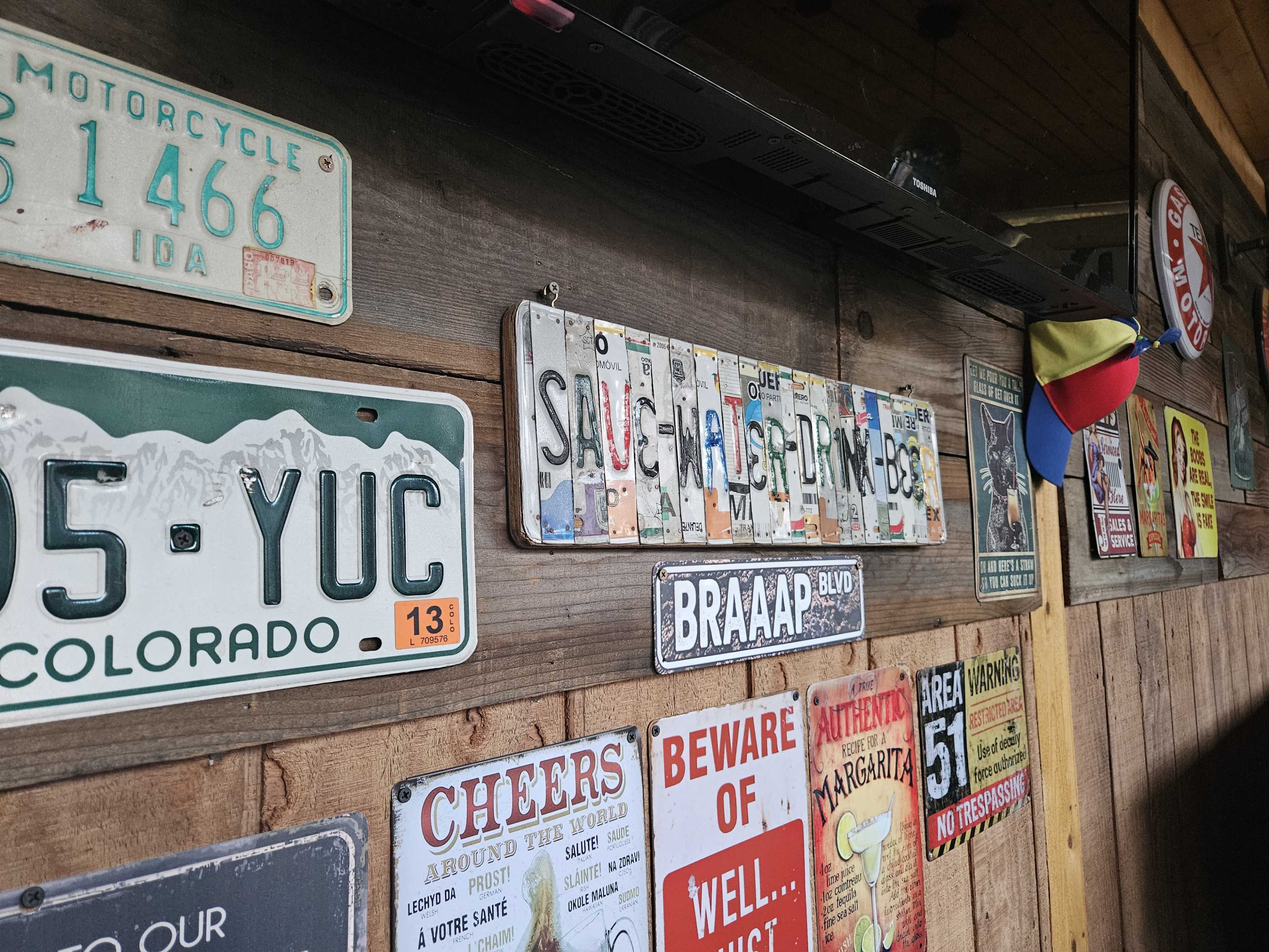 A wooden wall decorated with various license plates and vintage signs, alongside a colorful cap hanging on top.