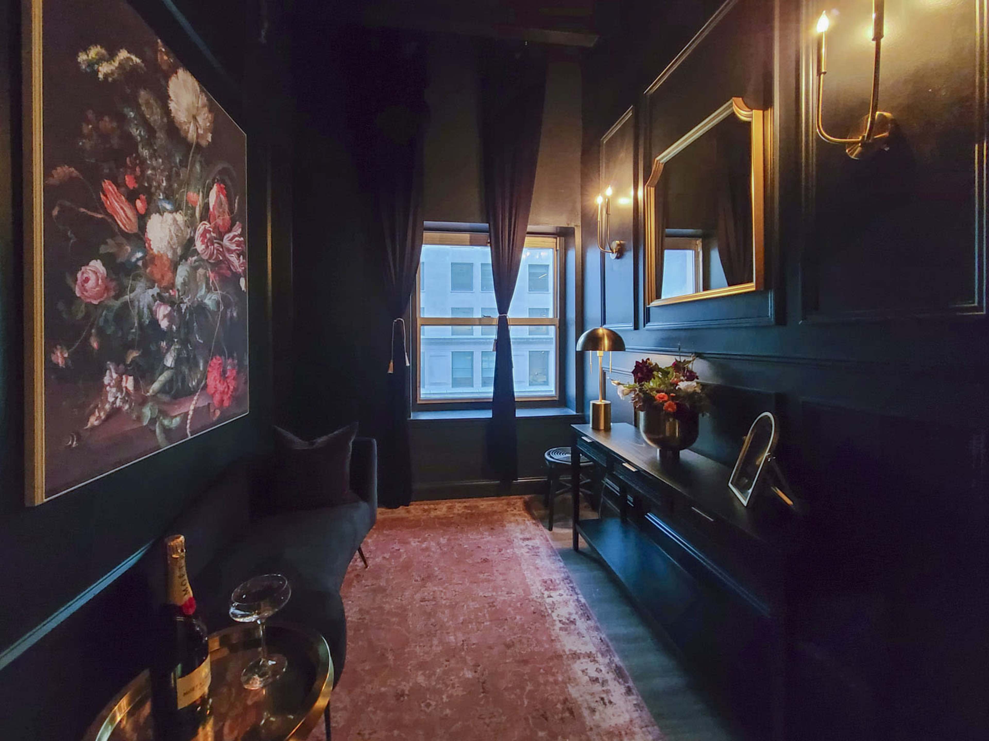 The image depicts a dimly lit, elegantly decorated corner of a room featuring a floral painting, a velvet bench, a console table with a lamp, and a window with dark curtains.