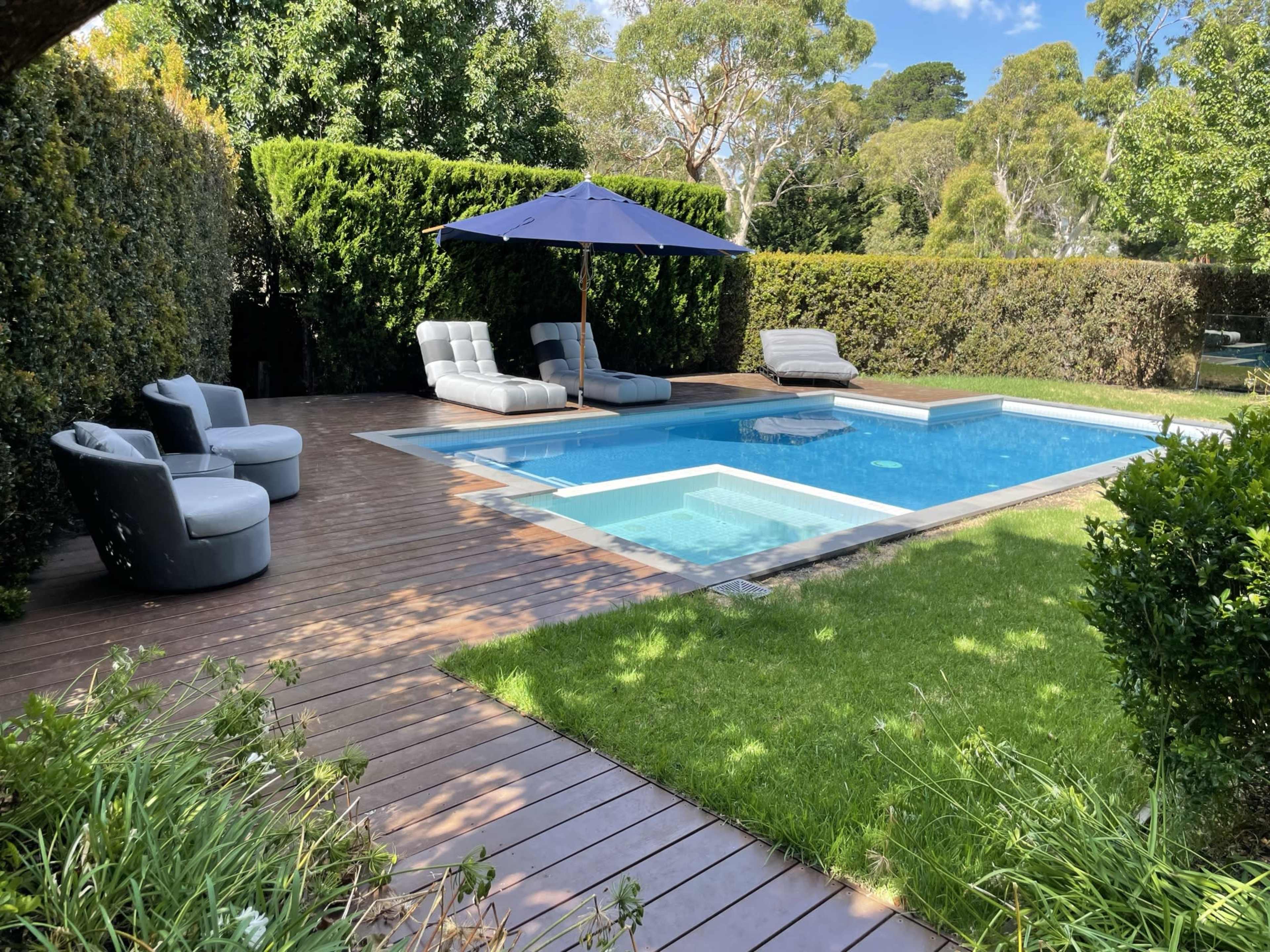 A well-maintained backyard features a swimming pool surrounded by sun loungers and an umbrella, with grass and wooden decking nearby.