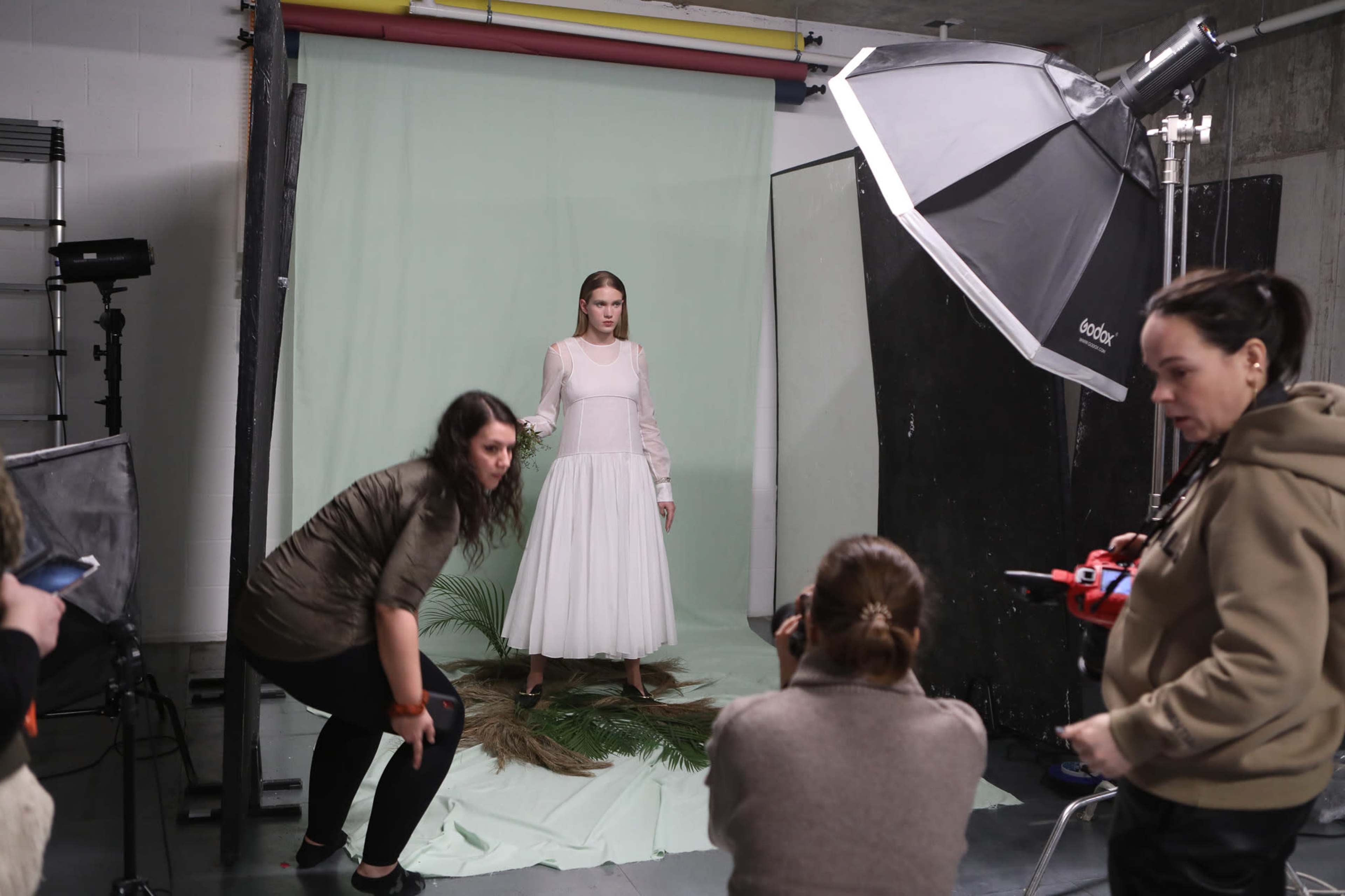 A model stands in a white dress on a green backdrop while two photographers adjust their equipment and take notes.