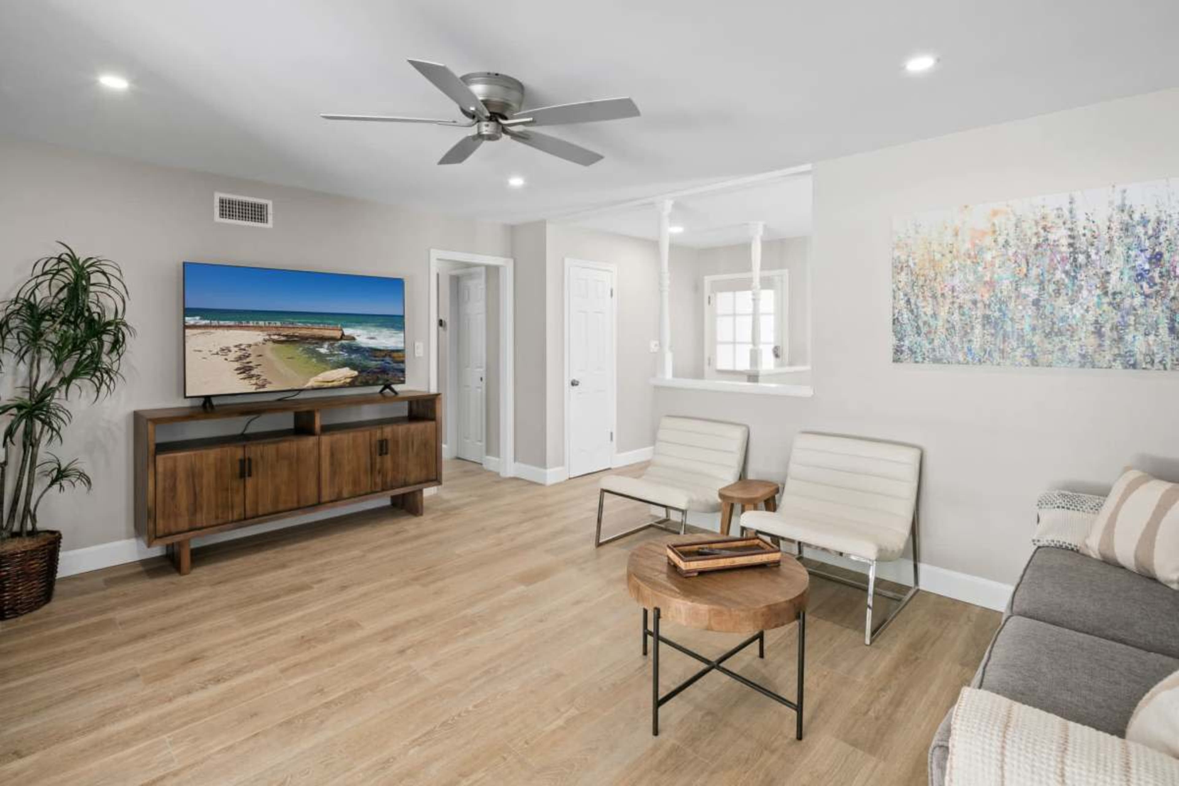 A modern living room features a television mounted on the wall, two white chairs, a wooden coffee table, and a gray sofa, with light wood flooring and neutral walls.