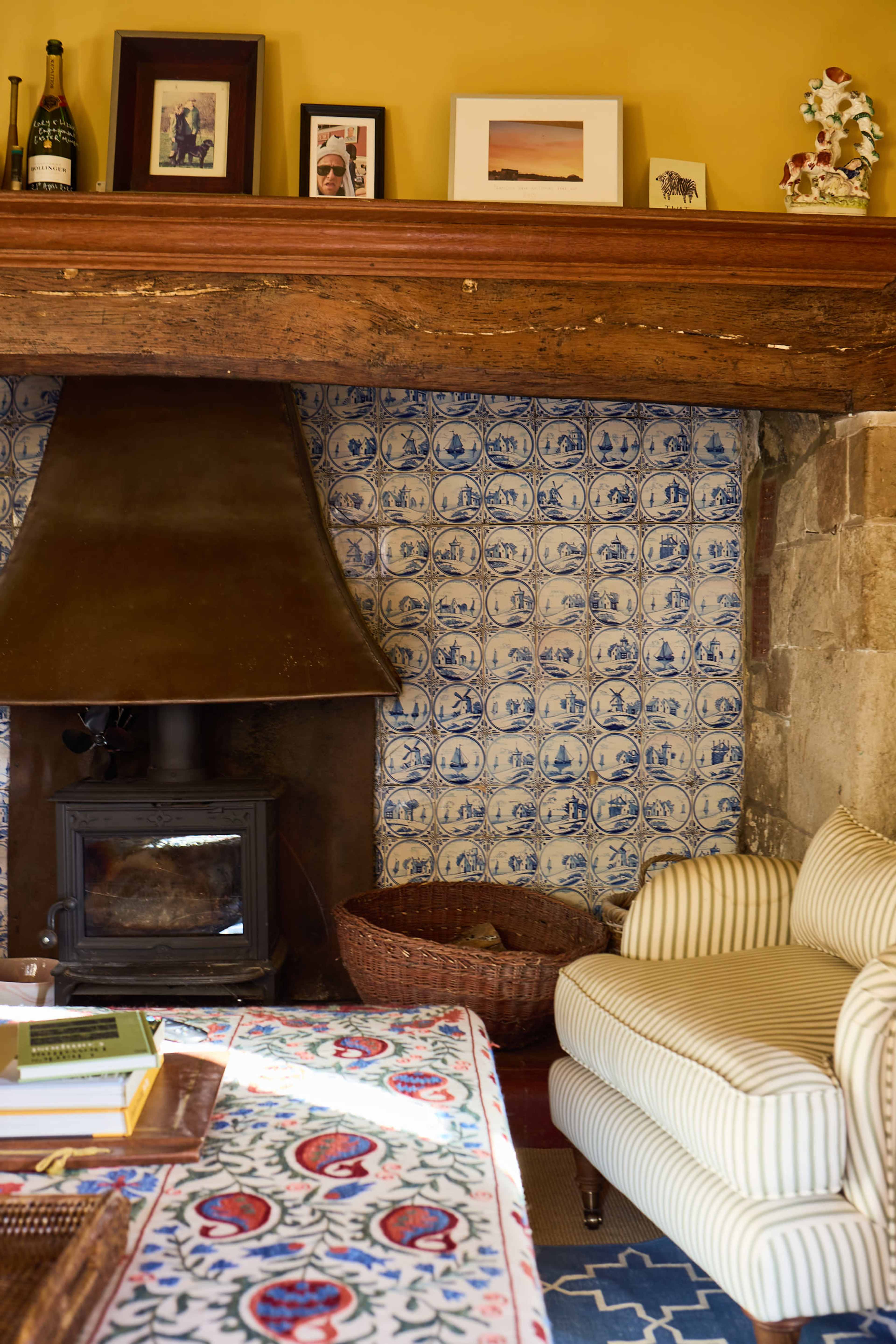 The image shows a cozy living room featuring a fireplace with a metal chimney, blue and white patterned tiles, a striped sofa, and a patterned tablecloth.