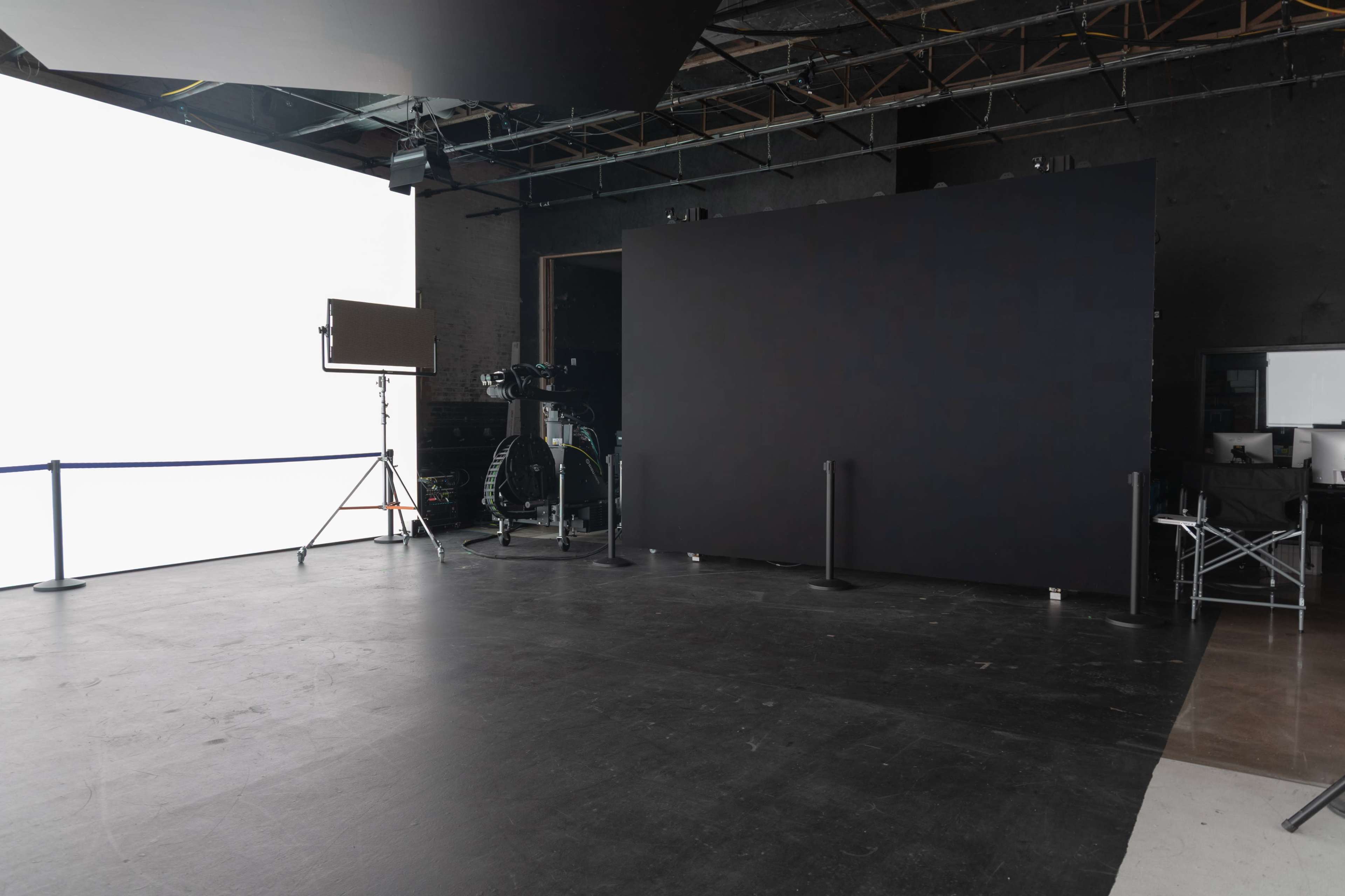 The image shows an empty studio space with a large black backdrop, a white light source, and a camera setup.