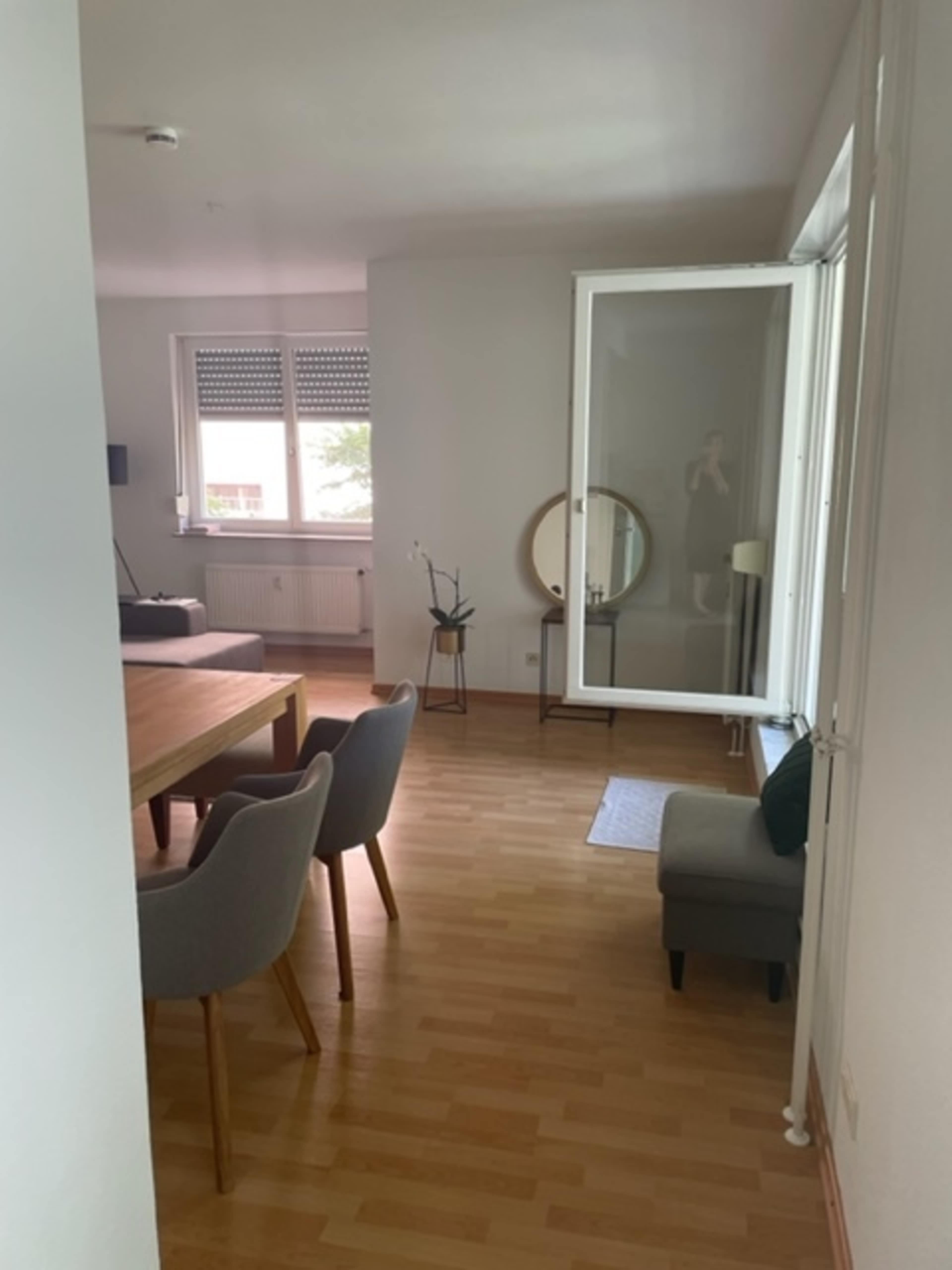 A bright, modern living space features a wooden dining table, gray chairs, and a large mirror next to a window.