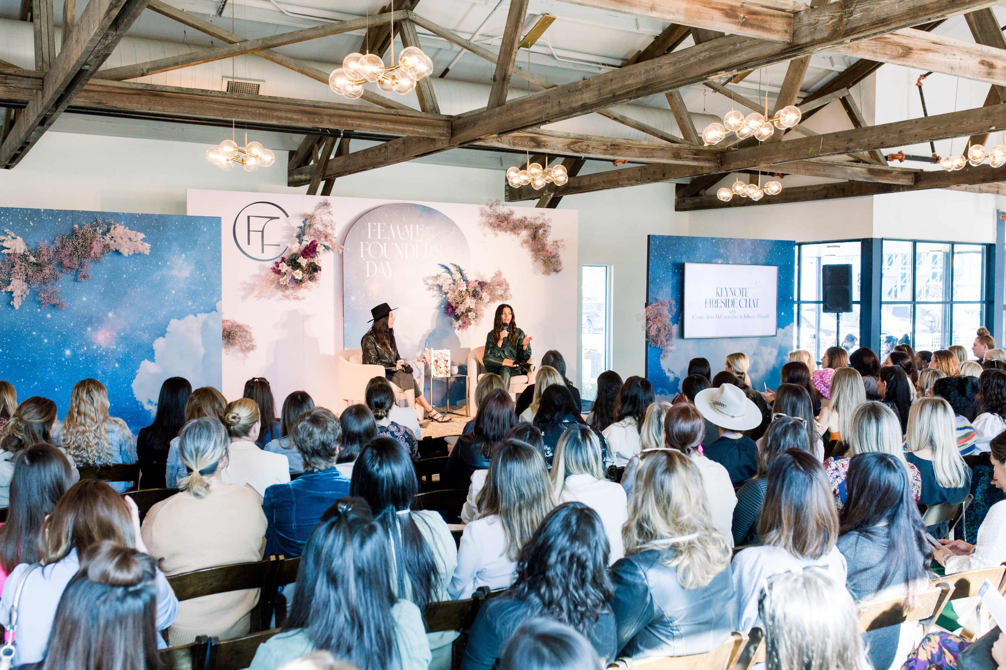 A large audience listens to a speaker at a panel event featuring a decorative stage with floral elements and branding.