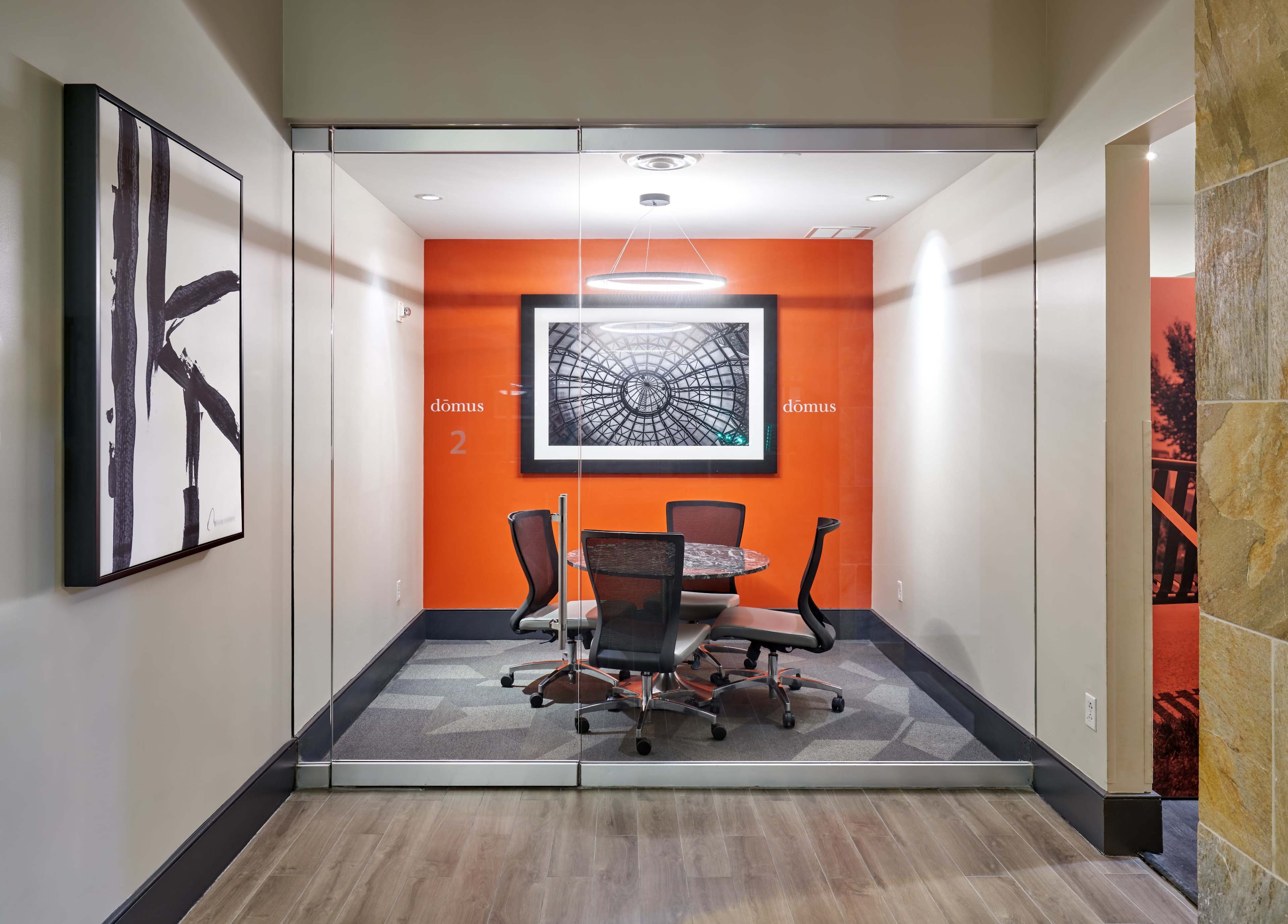 The image shows a modern conference room with a round table and four black chairs, enclosed by glass walls and featuring an orange accent wall.