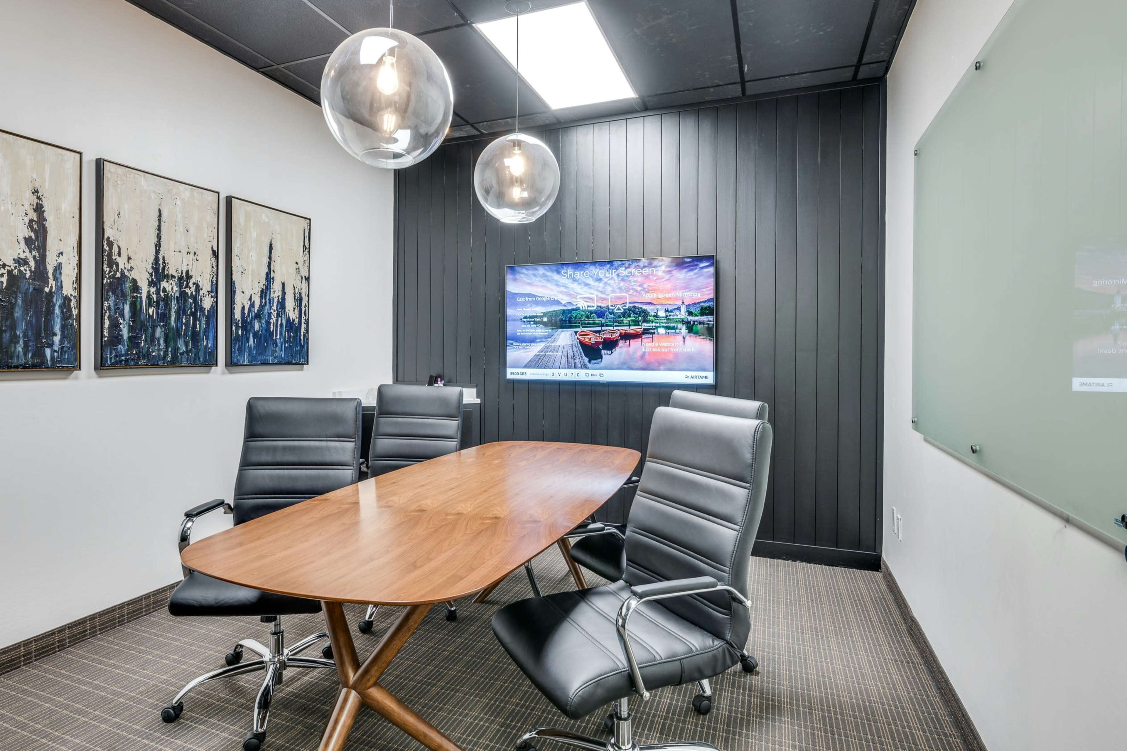 A modern conference room features a wooden table surrounded by black leather chairs, a large wall-mounted TV, and three hanging light fixtures.