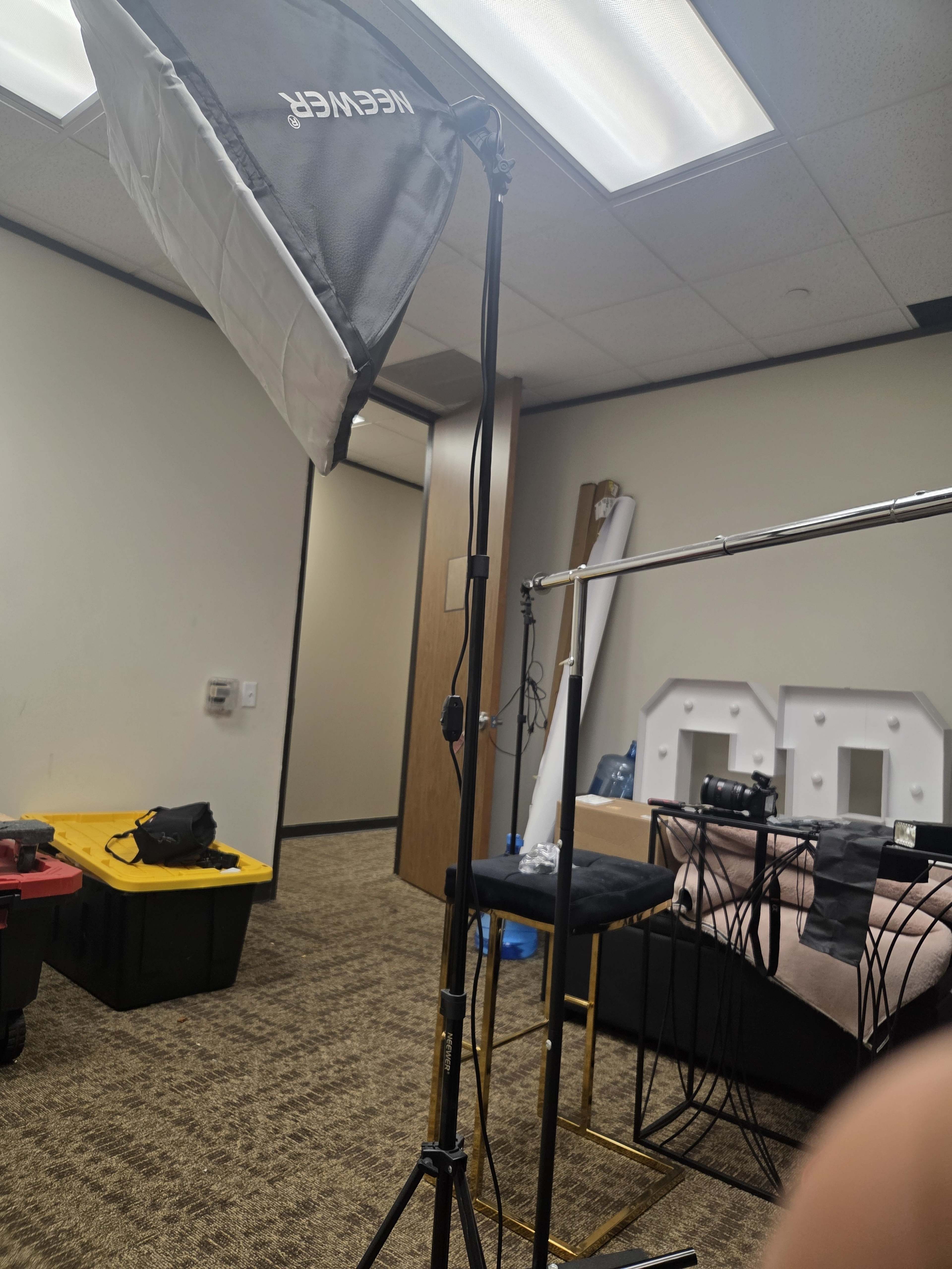 The image shows a photography setup in an office-like space, featuring a softbox light on a tripod and a clothing rack with various items.