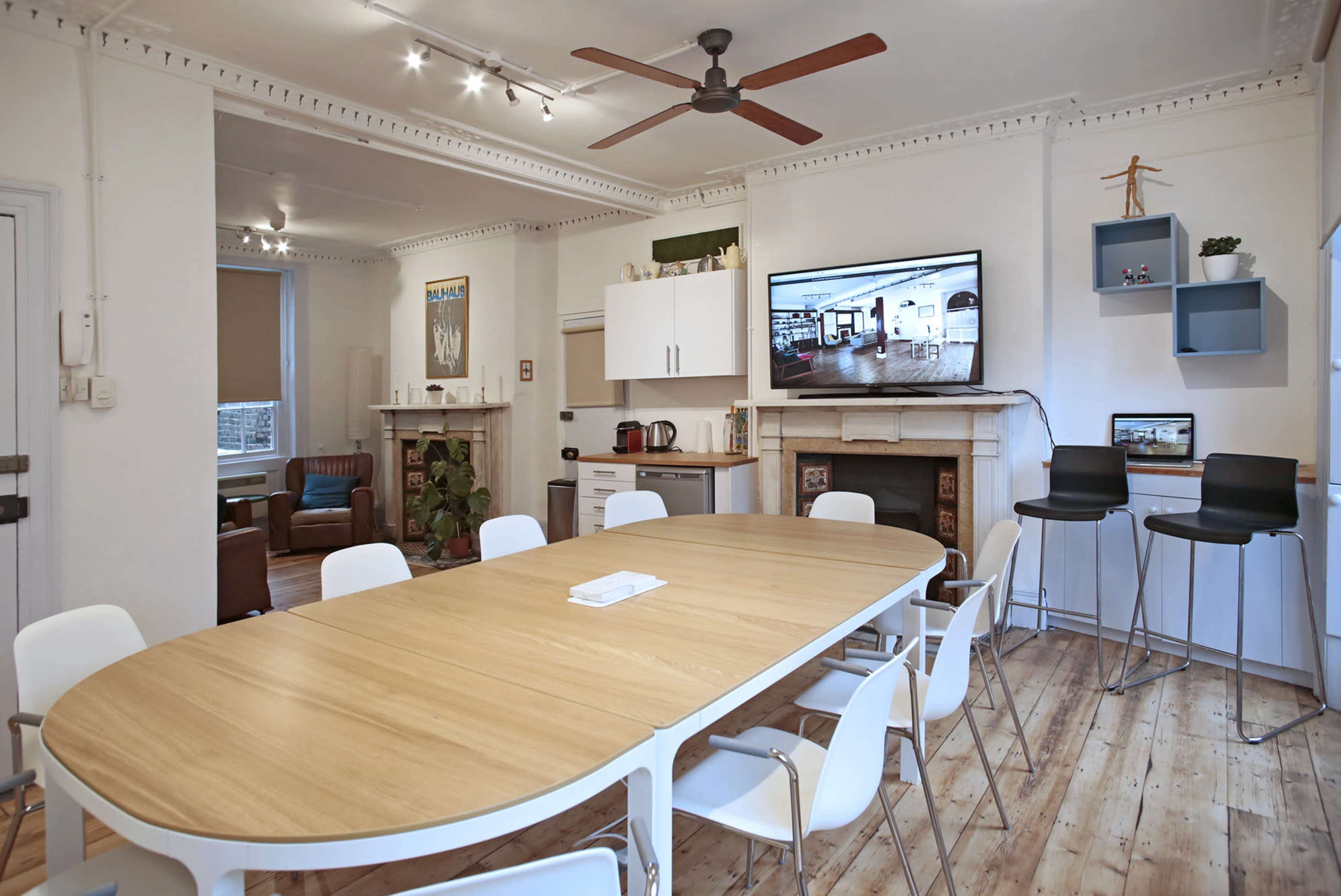 A spacious room features a wooden dining table surrounded by white chairs, with a television mounted on the wall and a kitchenette in the background.