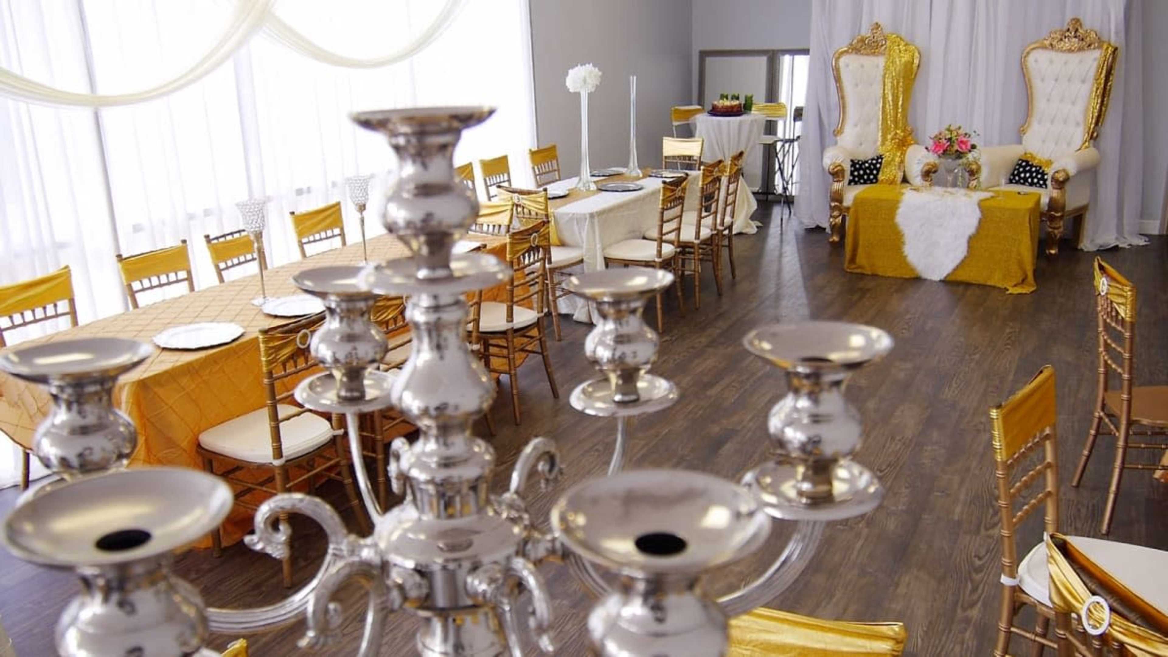 A decorated event space features golden chairs and tables with orange and white tablecloths, large white curtains, and elegant throne-like chairs at the front.