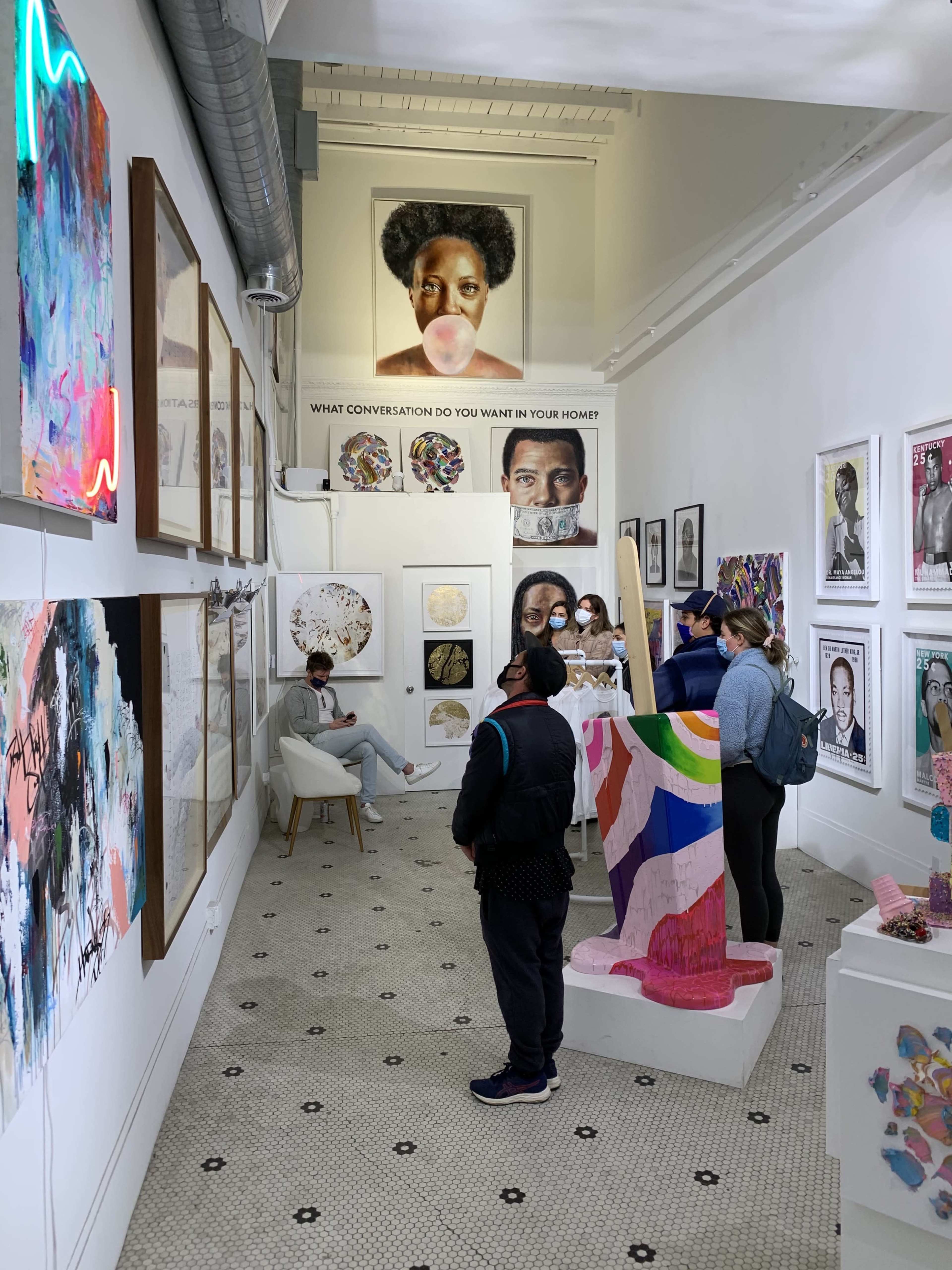 A group of people observes various artworks displayed on the walls of a gallery, with a large portrait and colorful sculptures prominently featured.