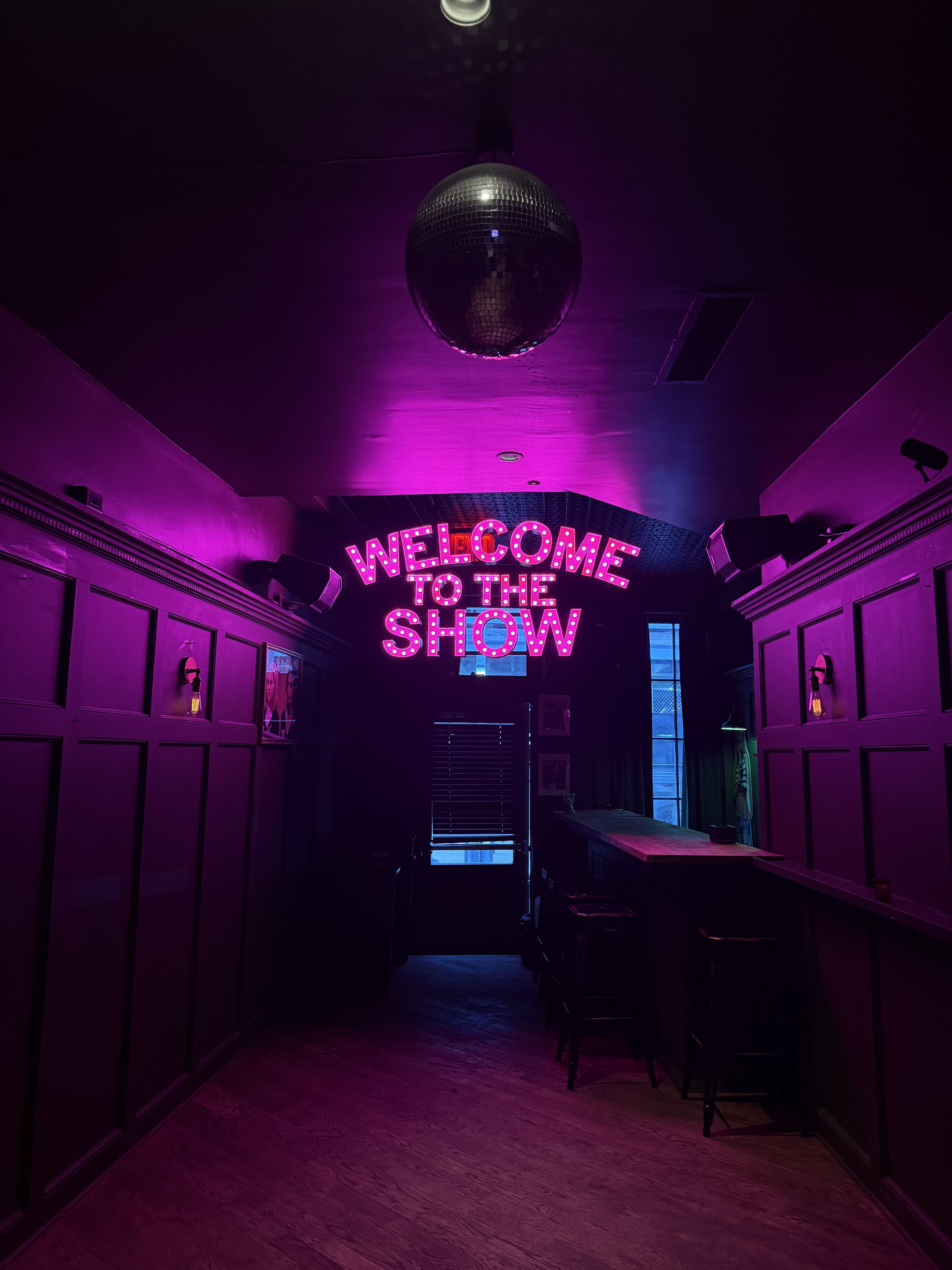 A dimly lit hallway features a neon sign that reads "WELCOME TO THE SHOW," with a disco ball hanging overhead.
