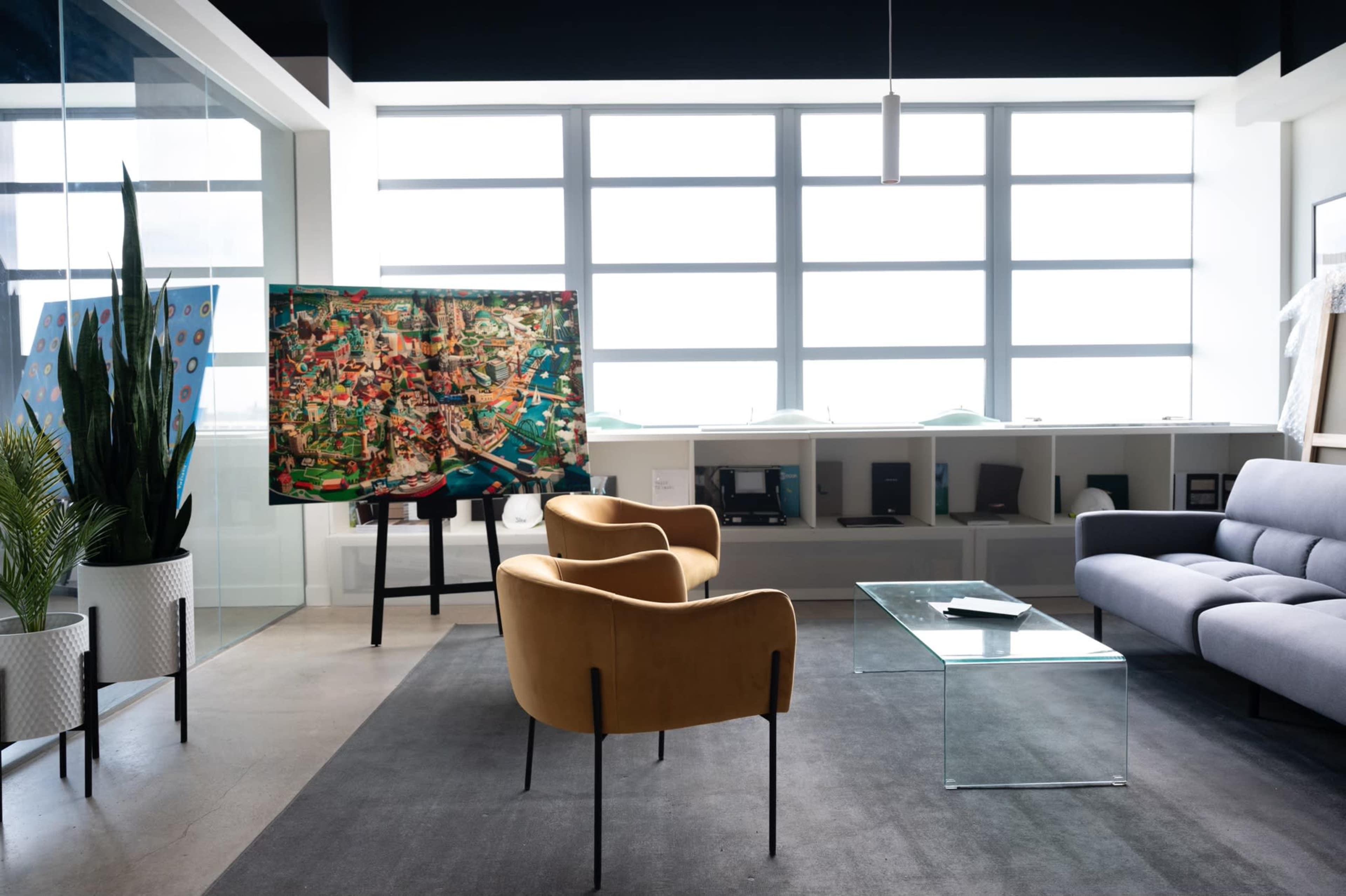A modern office space featuring two brown chairs, a glass coffee table, and a large colorful artwork displayed on an easel near large windows.