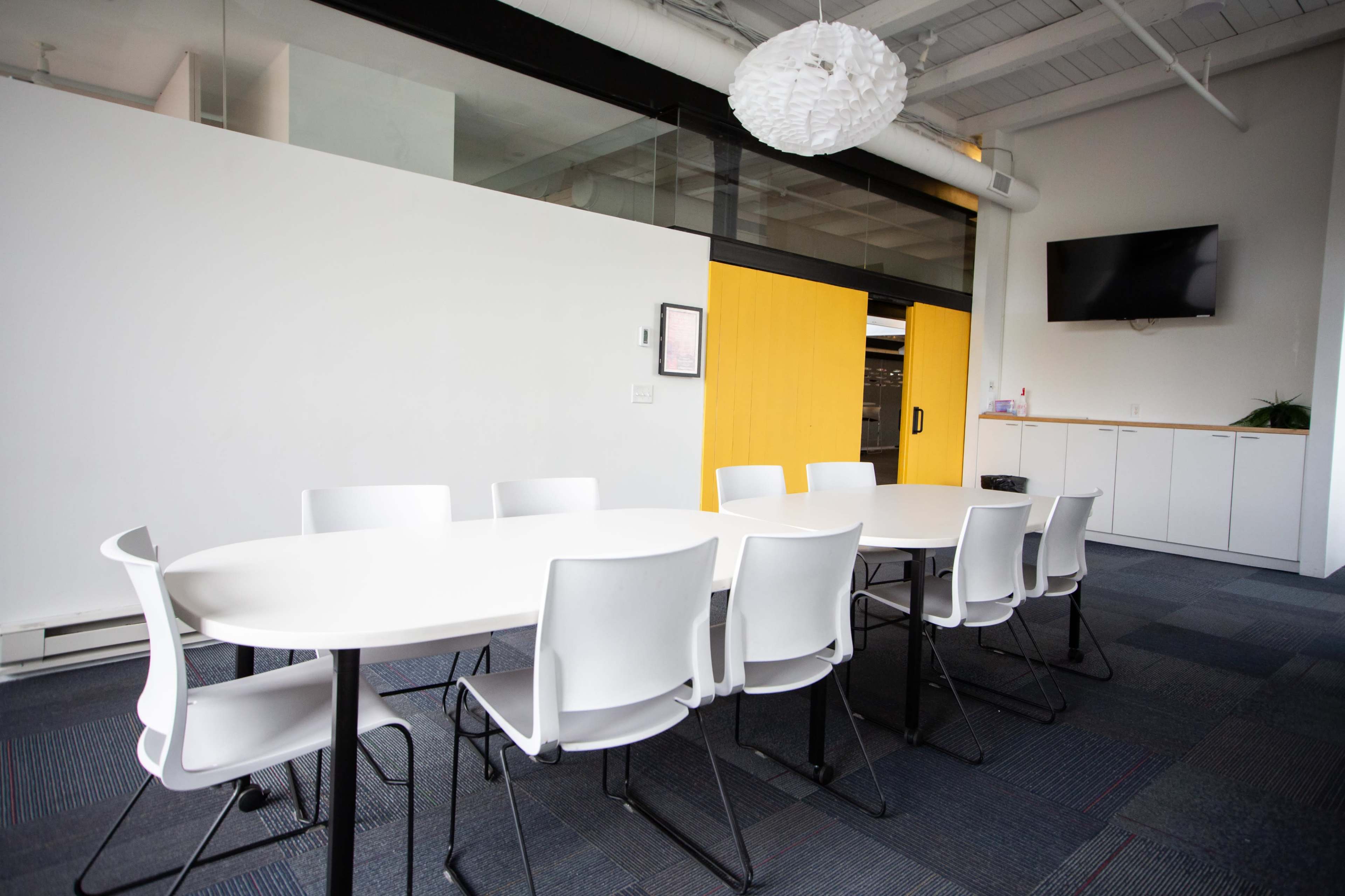 The image shows a modern meeting room with a round white table surrounded by white chairs, a TV mounted on the wall, and large glass windows.