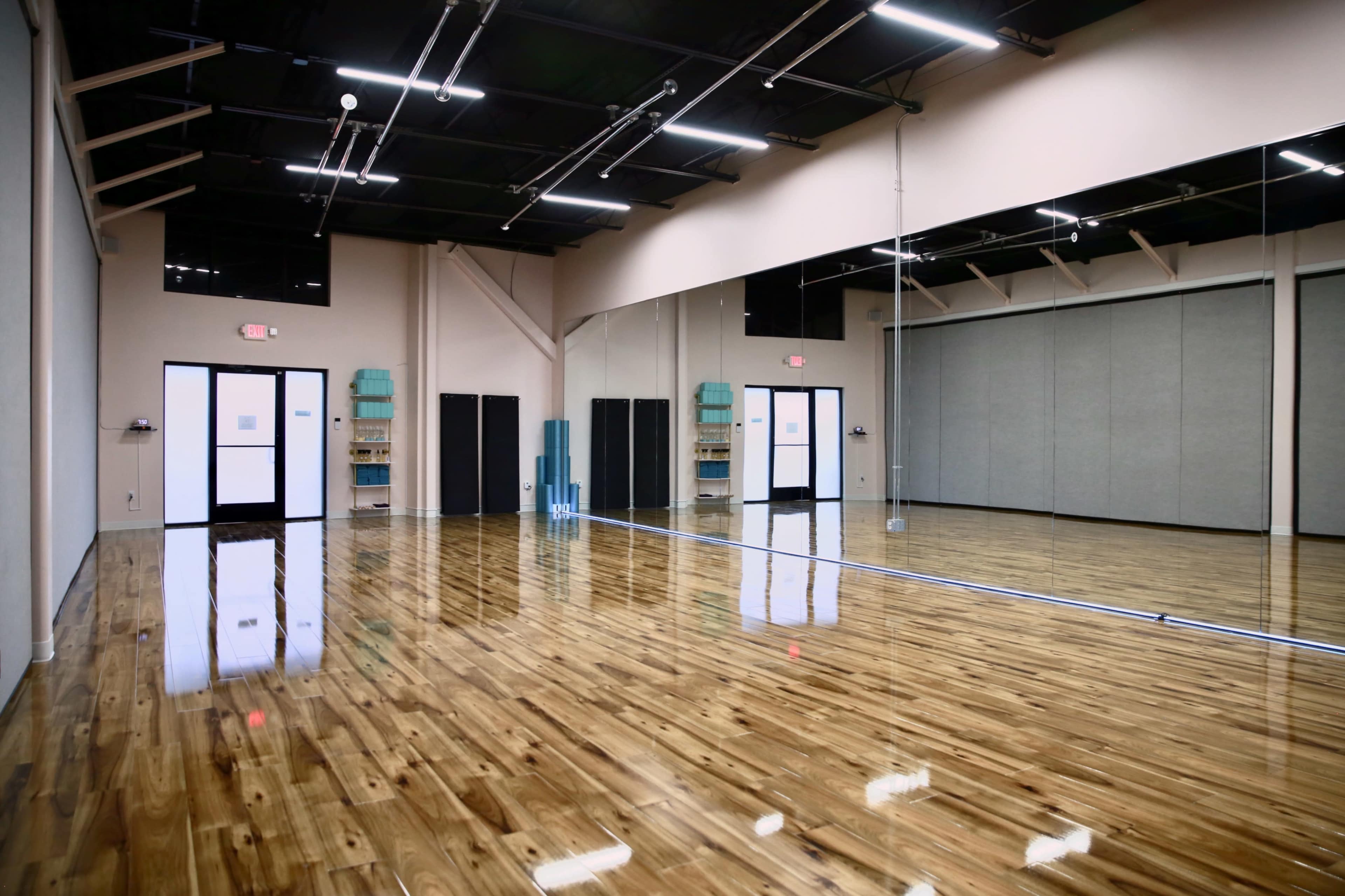 The image shows a spacious dance studio with polished wooden floors, large mirrors on one wall, and two doors leading to the outside.