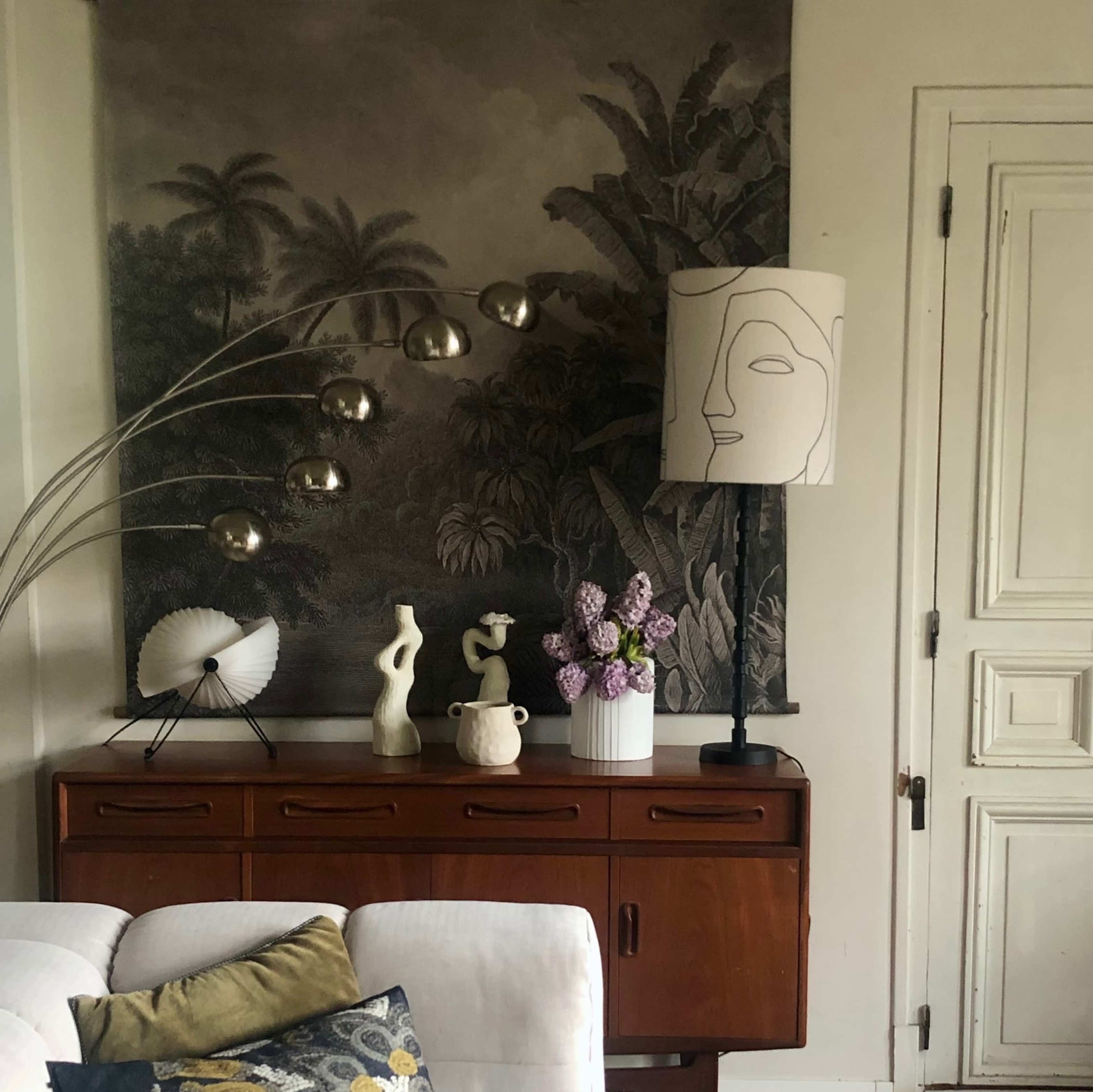 A mid-century sideboard displays decorative objects, a lamp, and a vase of flowers against a textured wall hanging.