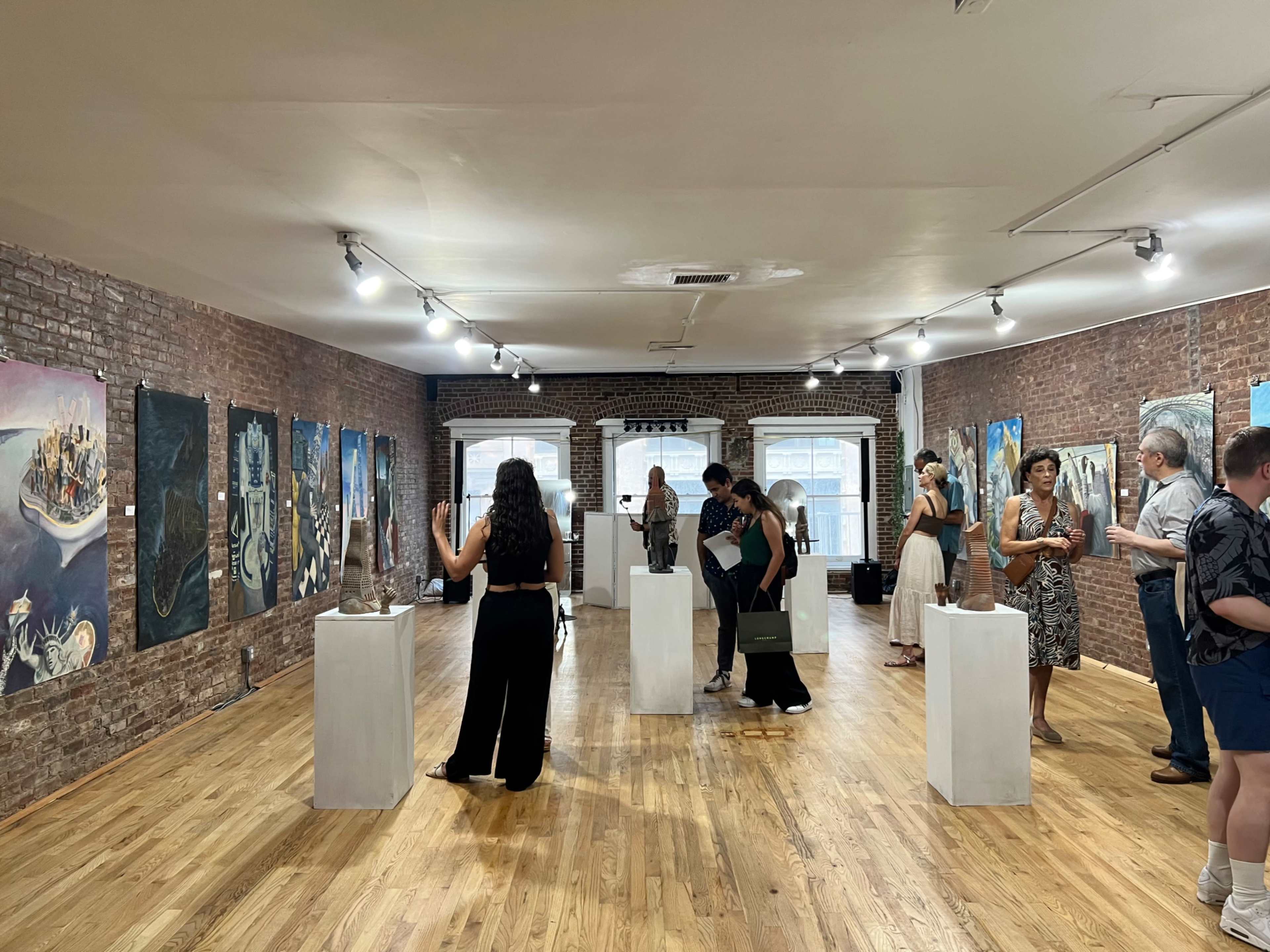 A group of people explore an art gallery featuring various paintings and sculptures displayed on pedestals in a room with wooden flooring and brick walls.