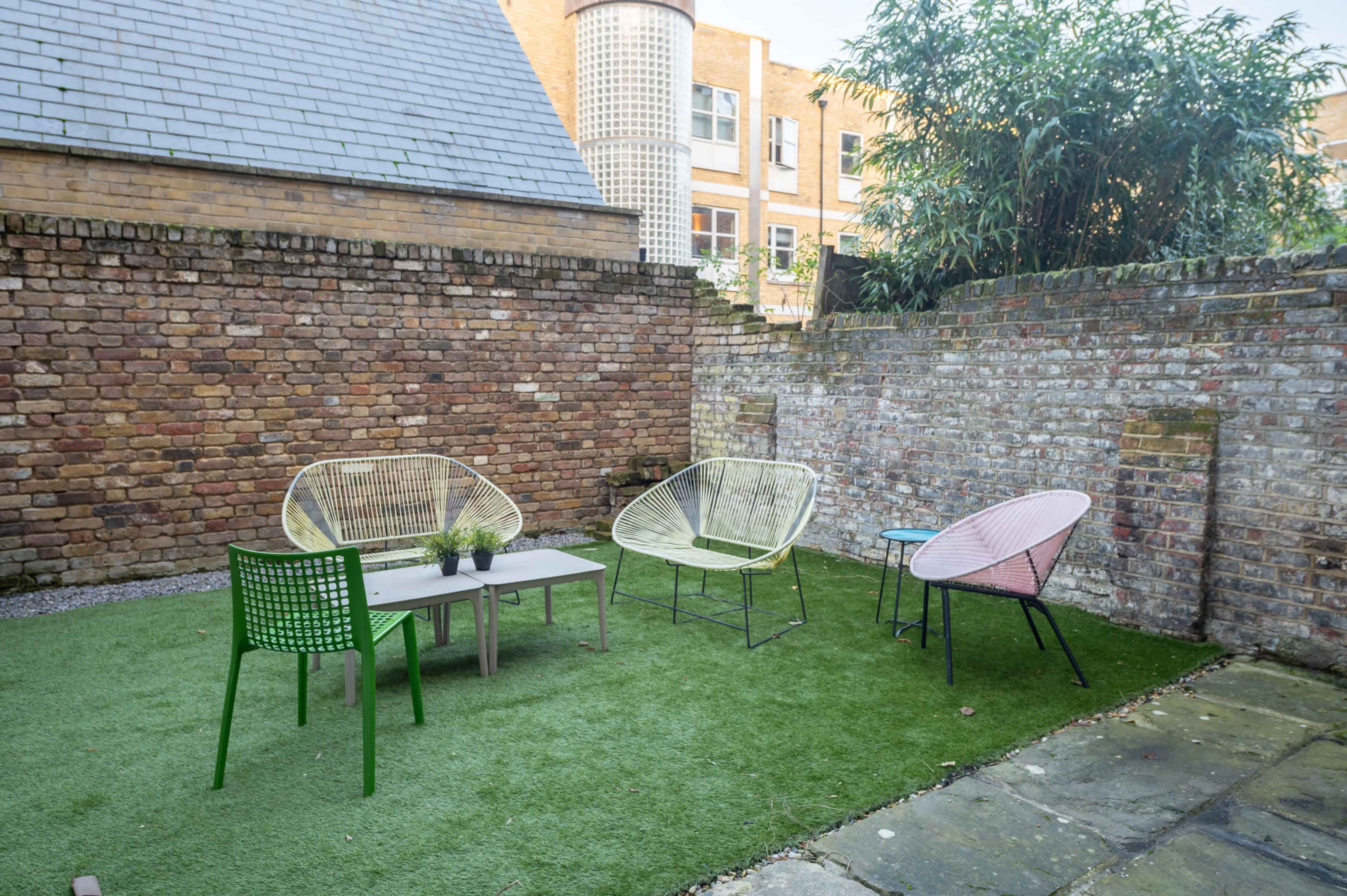 The image shows a small outdoor patio area with four modern chairs, a table, and a patch of artificial grass, surrounded by brick walls.