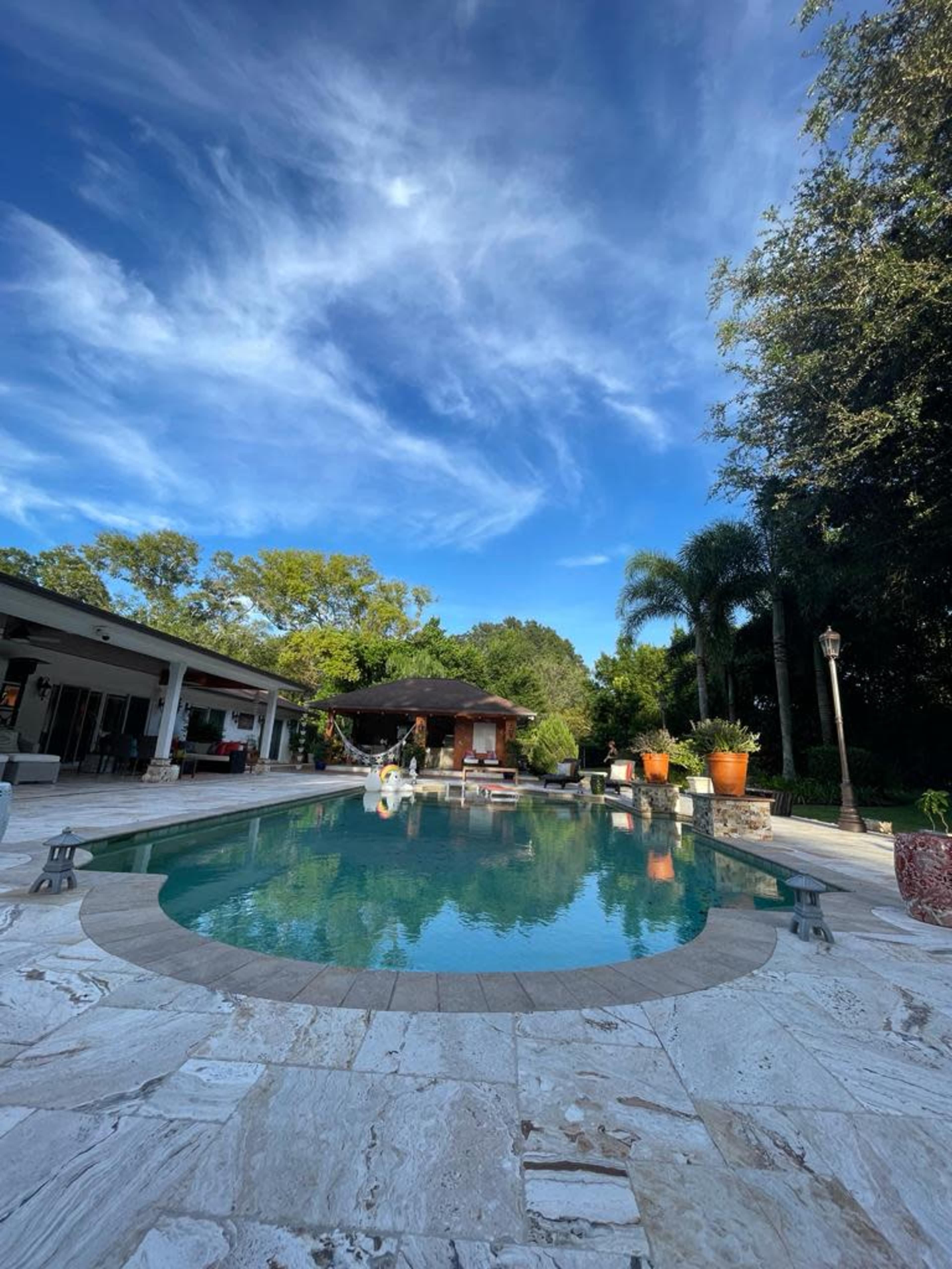 The image shows a swimming pool surrounded by lush greenery and a spacious outdoor area with a covered pavilion.