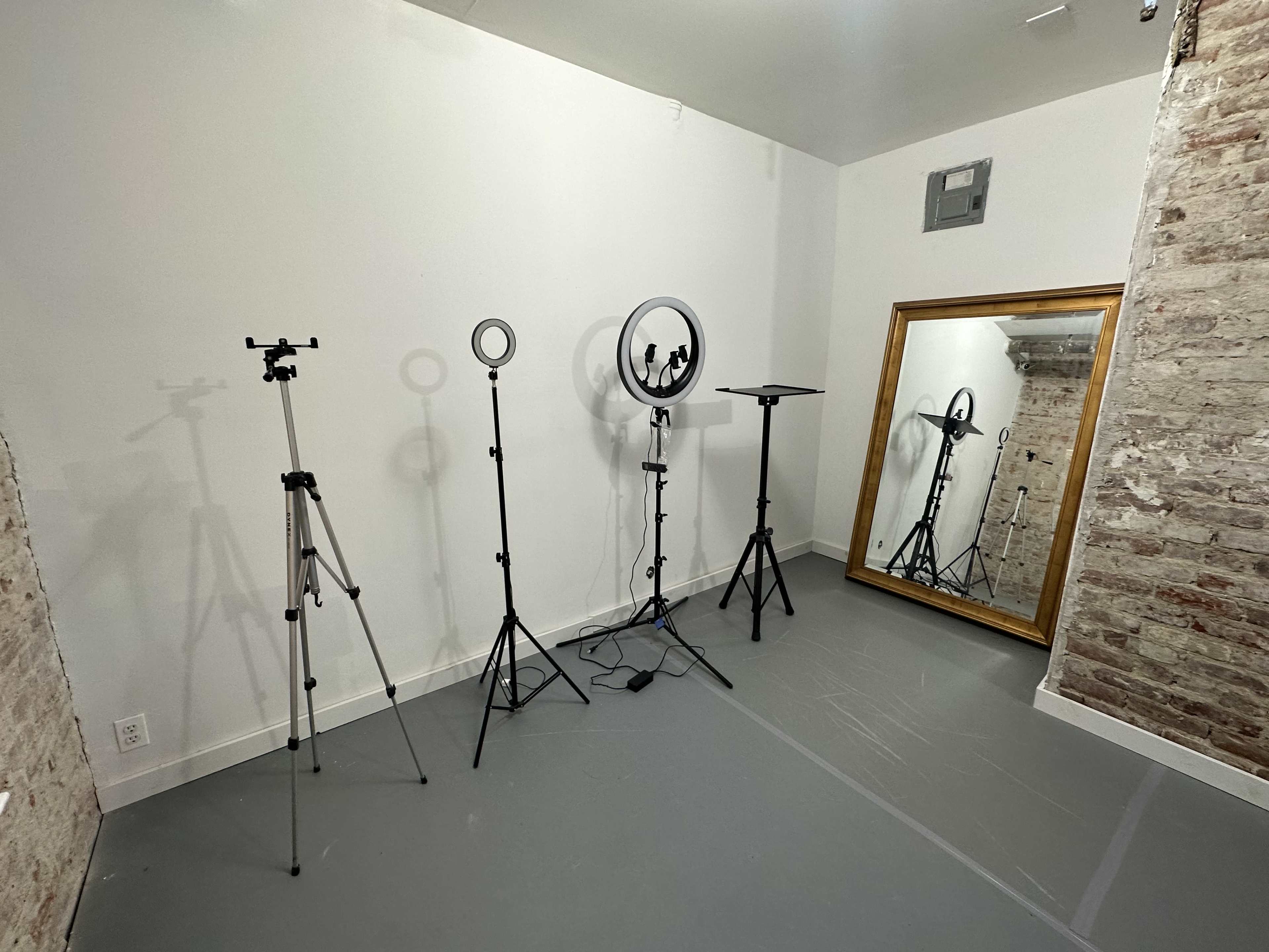 A minimalist room with a gray floor, featuring three tripods and a ring light, along with a large mirror against a brick wall.