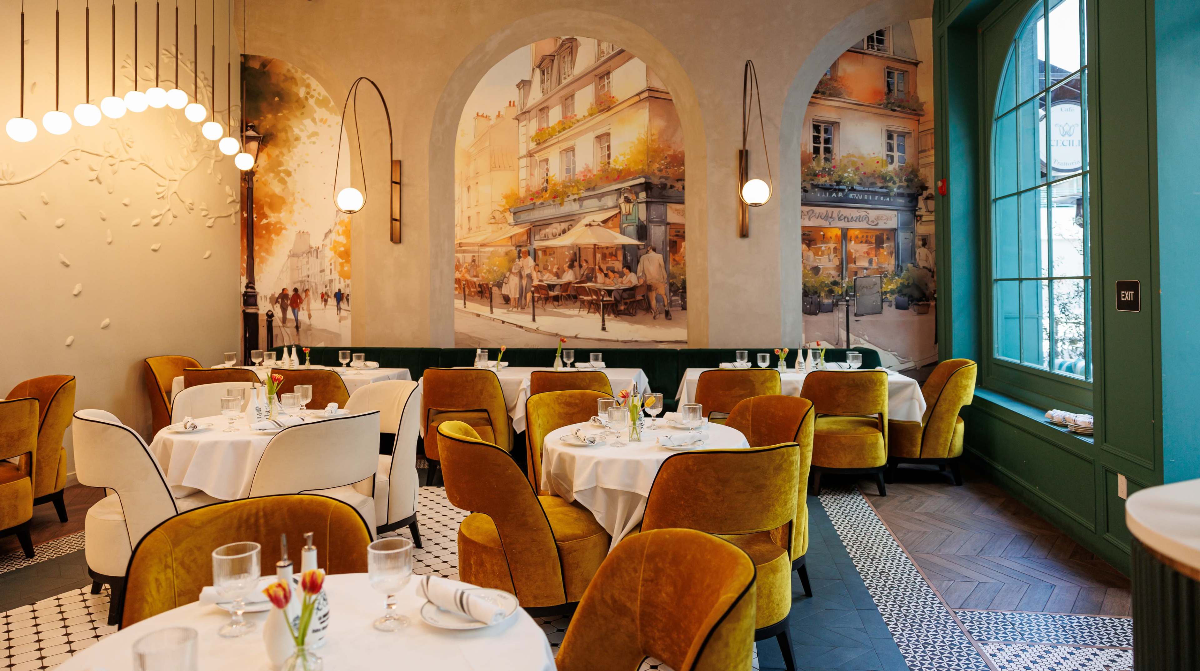 The image shows a restaurant interior featuring round tables with white tablecloths and mustard-colored chairs, adorned with wall murals of a street scene.