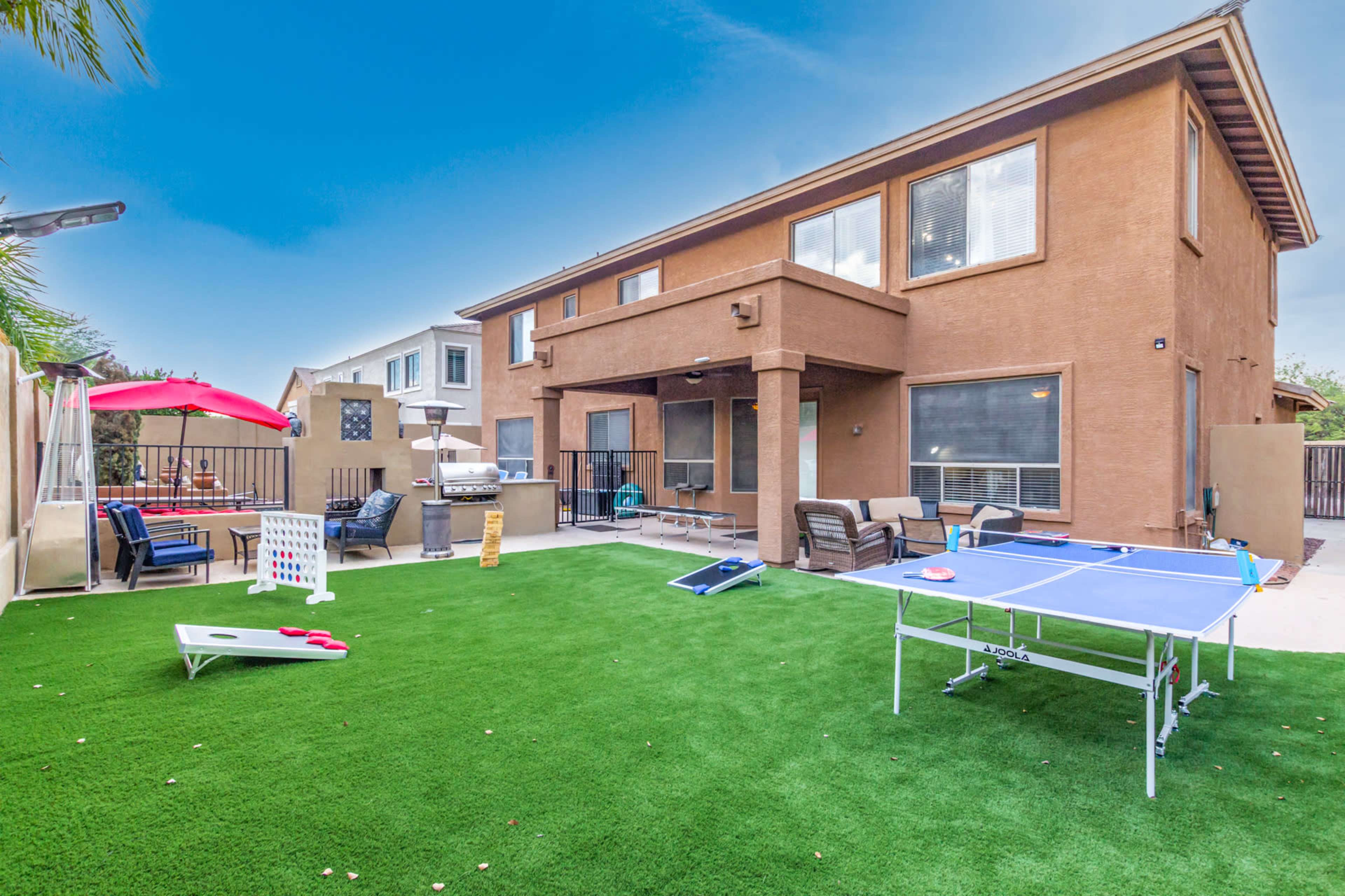 The image shows a backyard with artificial grass featuring a ping pong table, cornhole game, and seating areas next to a two-story residential home.