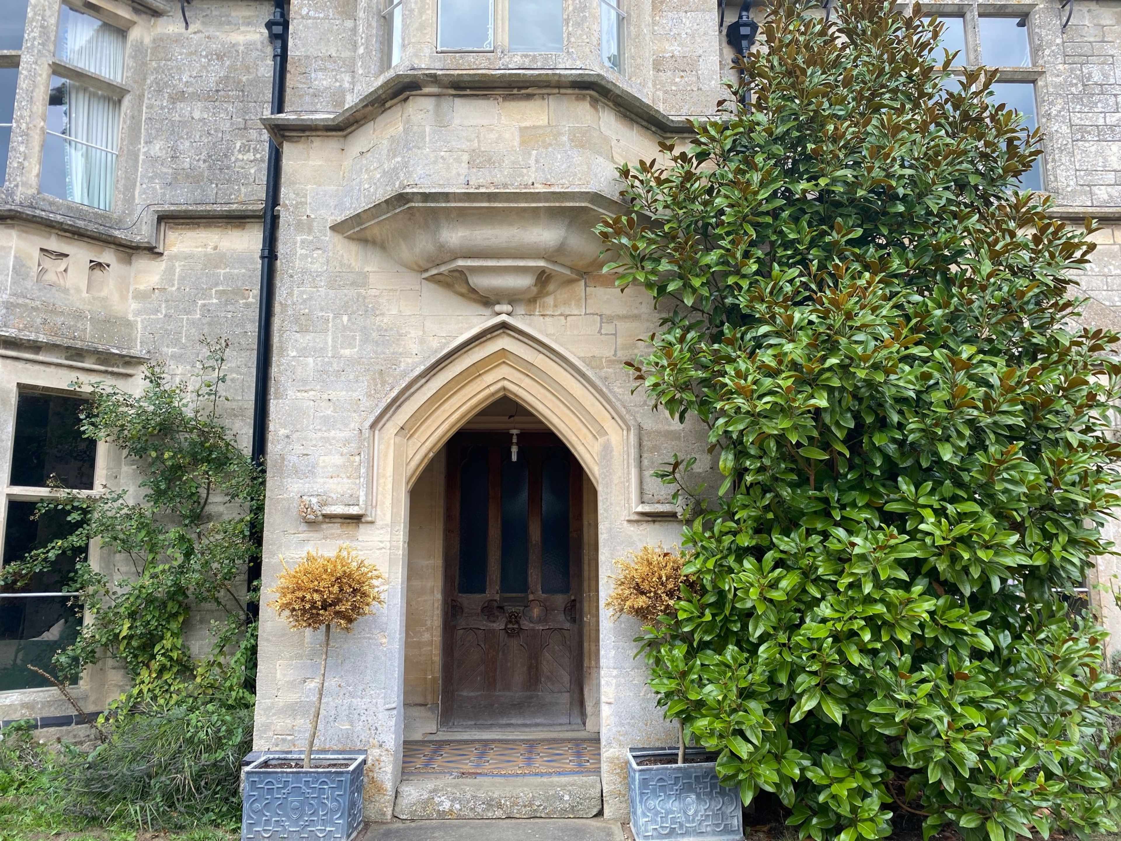 The entrance of a stone building with an arched wooden door, flanked by potted topiary trees.