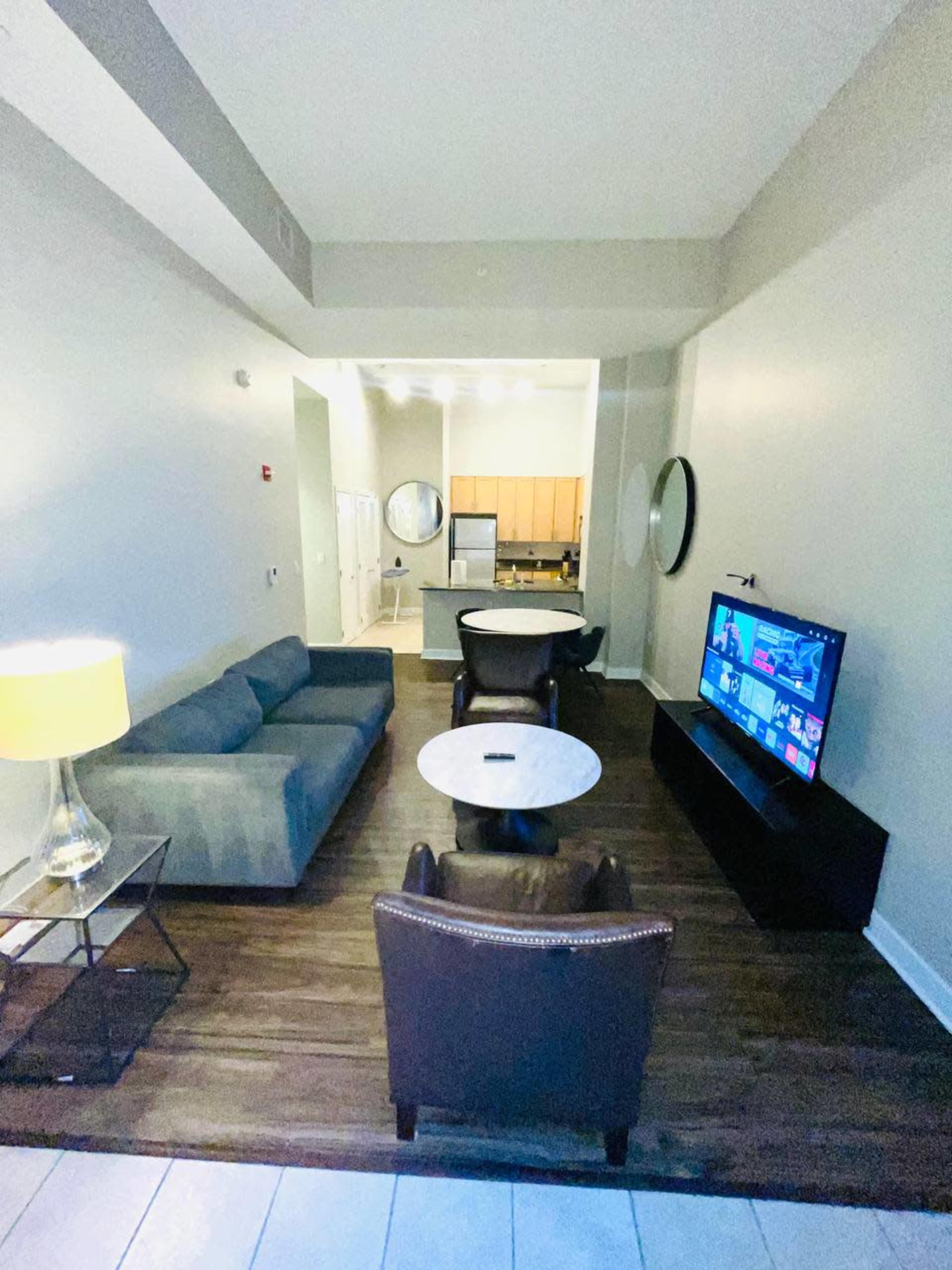 A modern living area with a gray sofa, two brown chairs, a round coffee table, a TV on a stand, and a lamp, set against a wall with a view of a kitchen in the background.