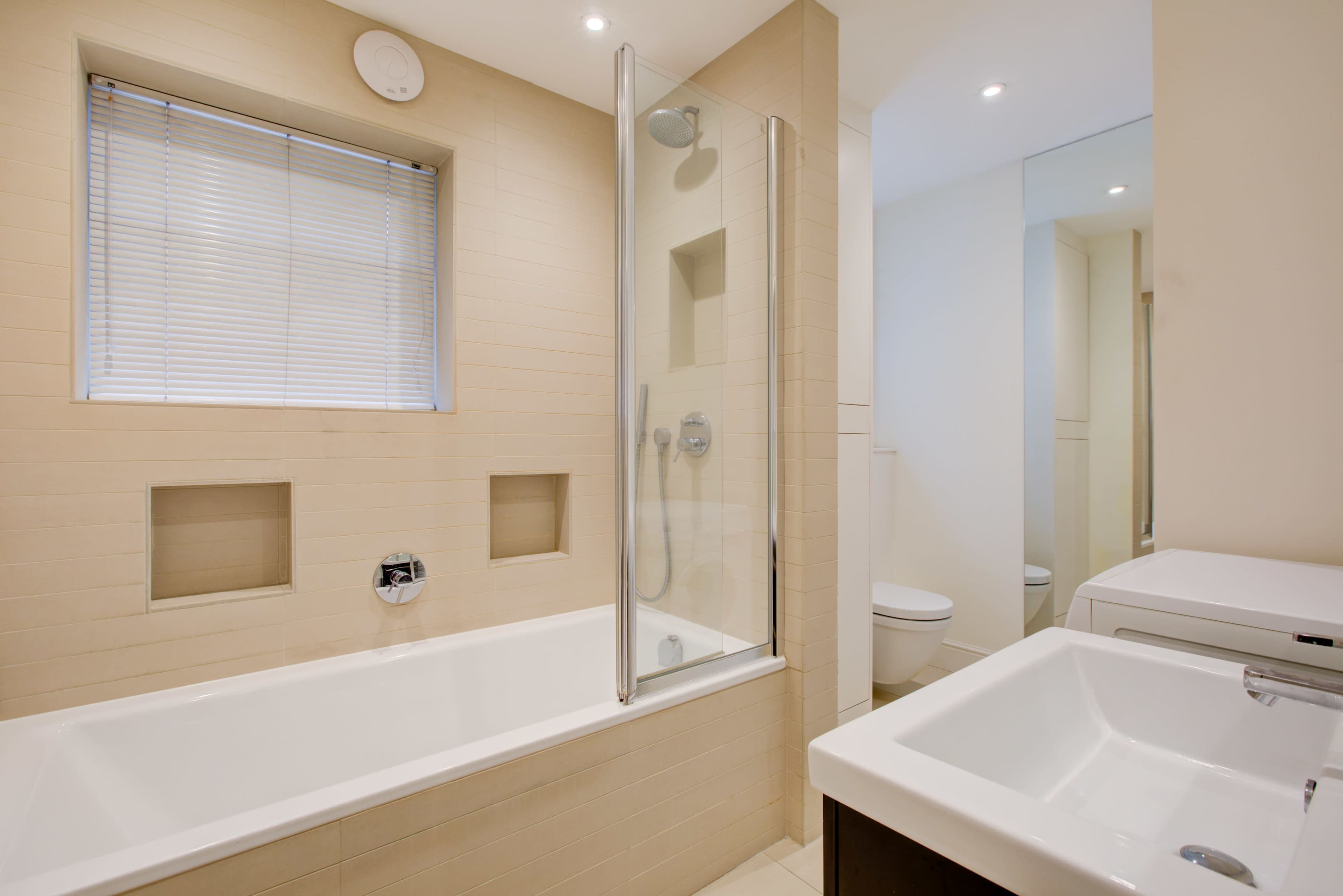 A modern bathroom featuring a bathtub with a glass shower enclosure, a sink, and a toilet, complemented by neutral-colored tile and a window with blinds.