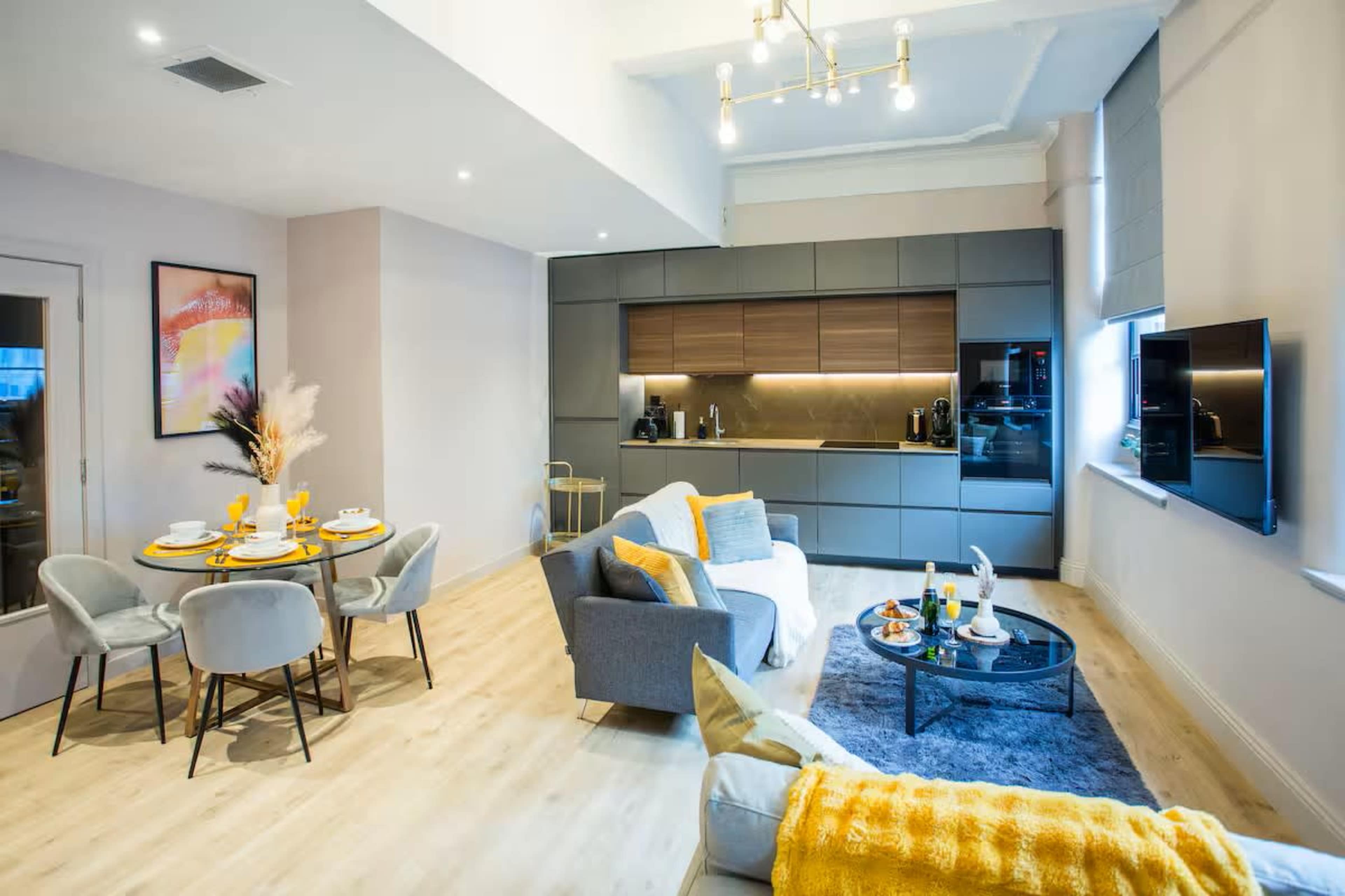 The image shows a modern living space featuring a dining area with a round table set for four, a gray sofa with yellow accents, and a sleek kitchen in the background.