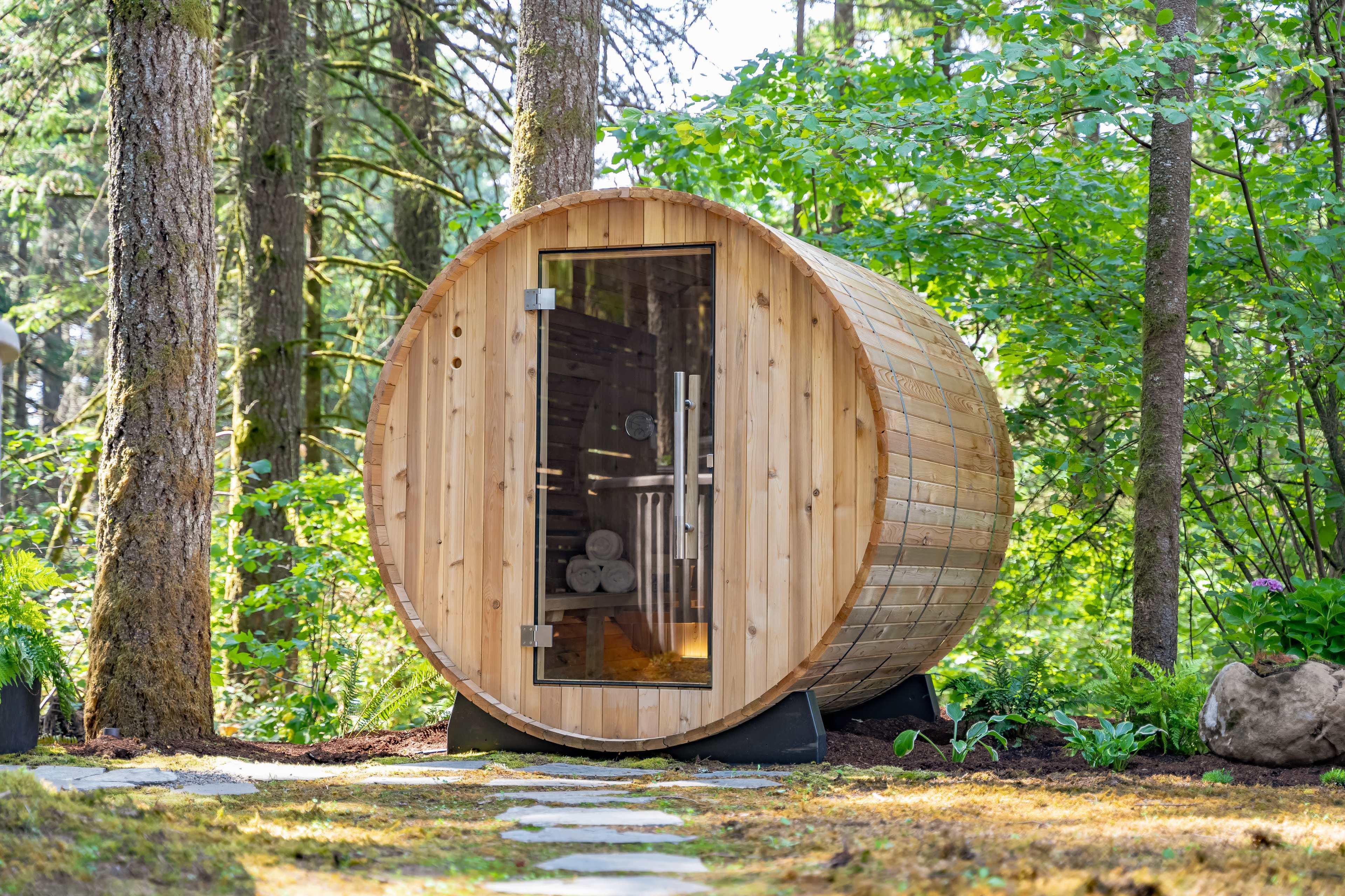 A wooden barrel sauna stands in a forested area surrounded by trees and greenery.